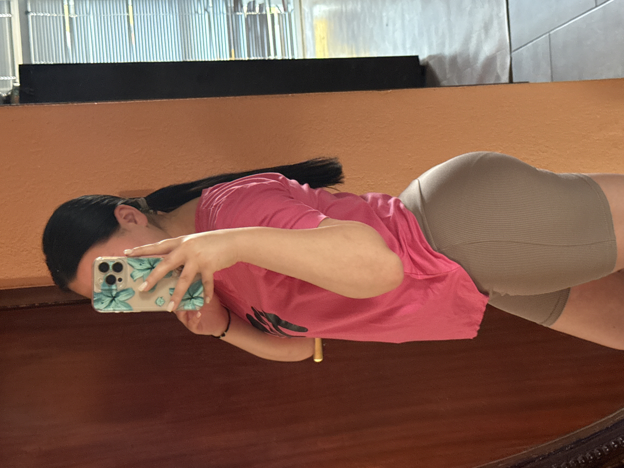 A person is depicted in an indoor setting in Rogosh, Bulgaria, taking a mirror selfie. The individual, seen from the left profile reflection, has dark hair tied into a ponytail and is holding a smartphone with their right hand, obscuring most of their face. The smartphone is an iPhone model, encased in a clear cover featuring a blue floral design, and the individual's fingernails are painted white. They are wearing a short-sleeved hot pink t-shirt with a dark, abstract logo on the left side of the chest, and form-fitting, ribbed, light brown or khaki bike shorts. A black beaded bracelet is visible on their left wrist. The immediate background to the right is an orange-brown textured wall. To the left, a darker brown surface, likely a door or frame, is reflected, featuring a horizontal golden handle. Further right, beyond the orange wall, a window with horizontal blinds is partially visible, showing an exterior or another room with a tiled floor. The overall lighting suggests an indoor environment with natural light entering from the window.