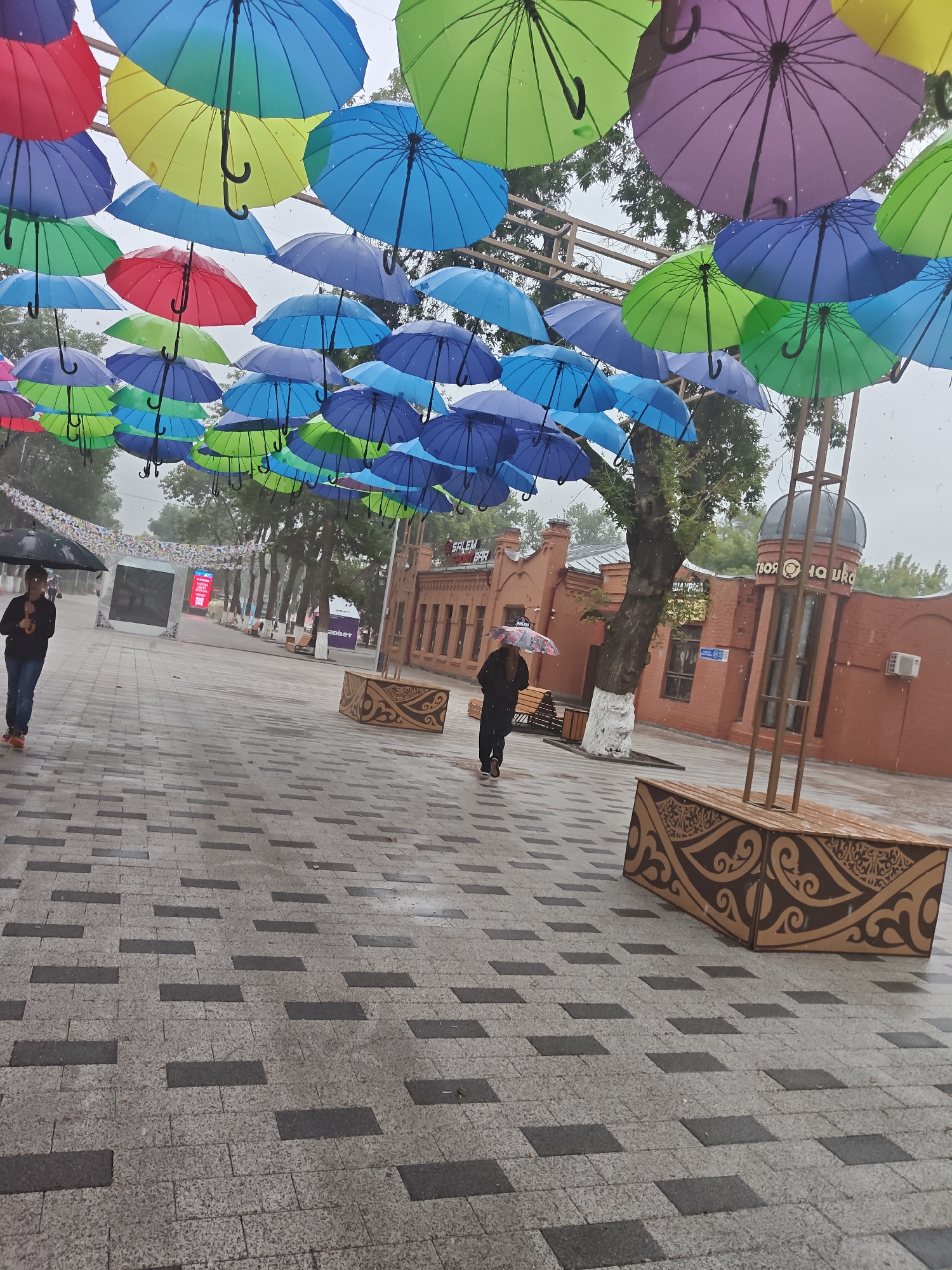 The image depicts an outdoor scene on a pedestrian walkway paved with interlocking stones. Above, a canopy of colorful umbrellas, predominantly in shades of blue, green, yellow, and red, is suspended, creating a vibrant overhead display. It appears to be raining, with visible water droplets in the air.
Two individuals are present on the walkway. In the foreground, a person walks towards the viewer's left, holding a black umbrella. Further down the walkway, another person is seen walking away from the viewer, holding a small umbrella with a colorful pattern.
In the background, there are brick buildings. One building, painted red, has several windows and an arched entrance. A sign with Cyrillic lettering, partially visible, is attached to one of the buildings. There is also a metal structure with a domed top. Several trees with green foliage are scattered throughout the scene.
Elements of urban infrastructure are visible, including a wooden bench with decorative carvings, streetlights, and what appears to be a digital display screen. The overall atmosphere suggests a public space in a city, likely a park or a street designed for pedestrian use, decorated for an event or season. The location is identified as Petropavl, Kazakhstan.