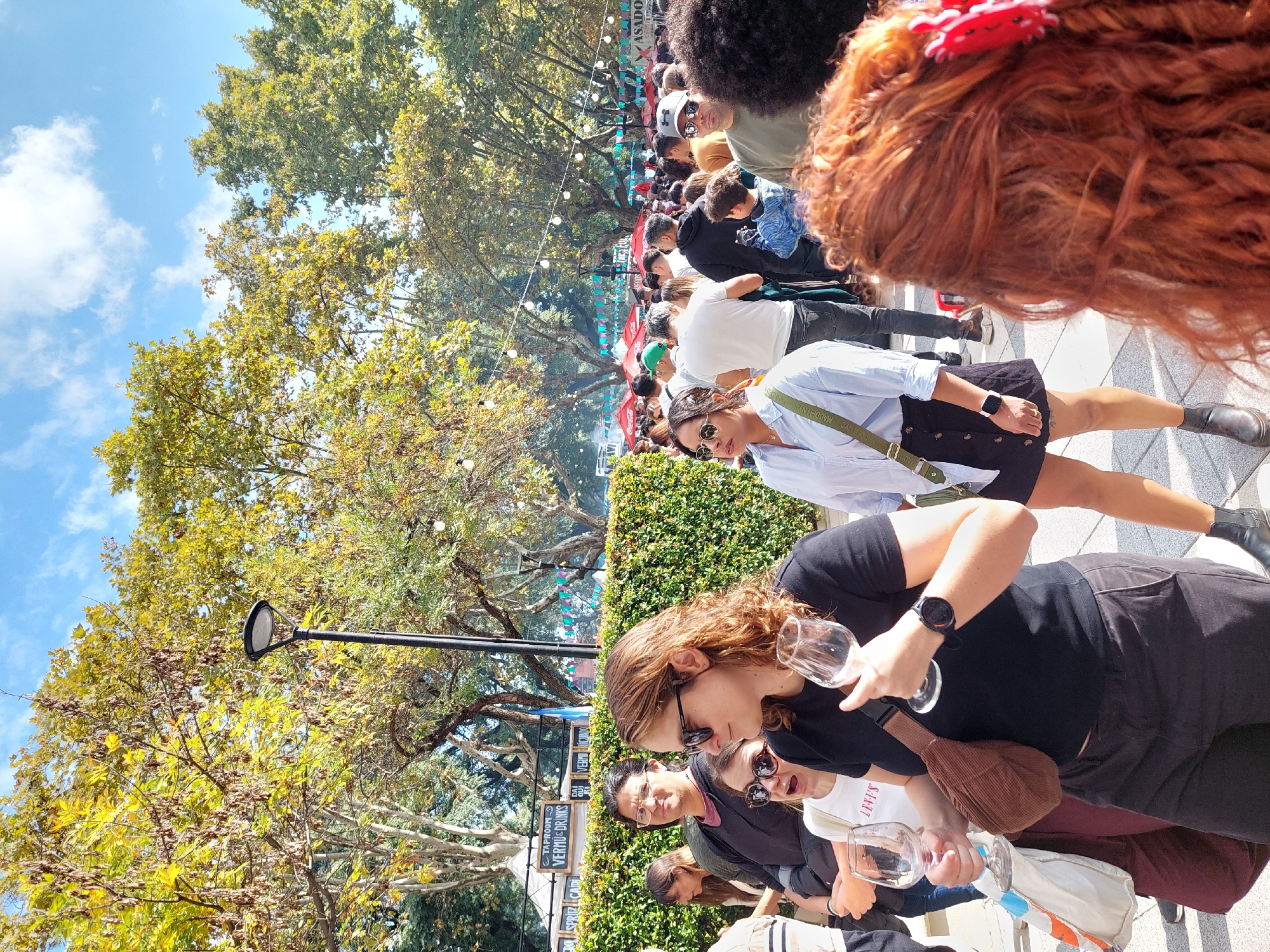A vibrant outdoor event is captured on a sunny day, bustling with numerous people enjoying a social gathering. The scene appears to be a street or park pathway lined with trees and market stalls, under a bright blue sky with scattered white clouds. String lights are strung across the path, adding to the festive atmosphere.
In the foreground, a woman with reddish-brown curly hair, wearing a black short-sleeved top, dark pants, and a black smartwatch, holds two empty wine glasses and a brown corduroy fanny pack. Beside her, another woman with brown hair and sunglasses wears a white t-shirt and also holds an empty wine glass. Slightly behind them, a woman in a light blue shirt, black skirt, and sunglasses has a bag strap visible with "MADDEN NYC" printed on it. A man with glasses and a dark shirt stands near them.
Further down the path, a diverse crowd of people, including adults and some children, are seen walking, standing, and socializing. Green hedges line the path, and tall trees with green and yellowing leaves provide shade. A lamppost is visible amidst the foliage.
The event seems to be a food and drink festival or market. Visible text on banners or stalls includes "TAPROOM", "VERYU DRINKS", and "SPRITZ CABA", indicating beverage offerings. The acronym "CABA" suggests the location is the Ciudad Autónoma de Buenos Aires, as confirmed by the provided context. In the distant background, "Asado" is faintly visible, hinting at Argentine barbecue, and a "Coca-Cola" logo can also be seen. People appear relaxed and engaged, indicating a pleasant social occasion. The time of day is daytime, with clear, bright weather.