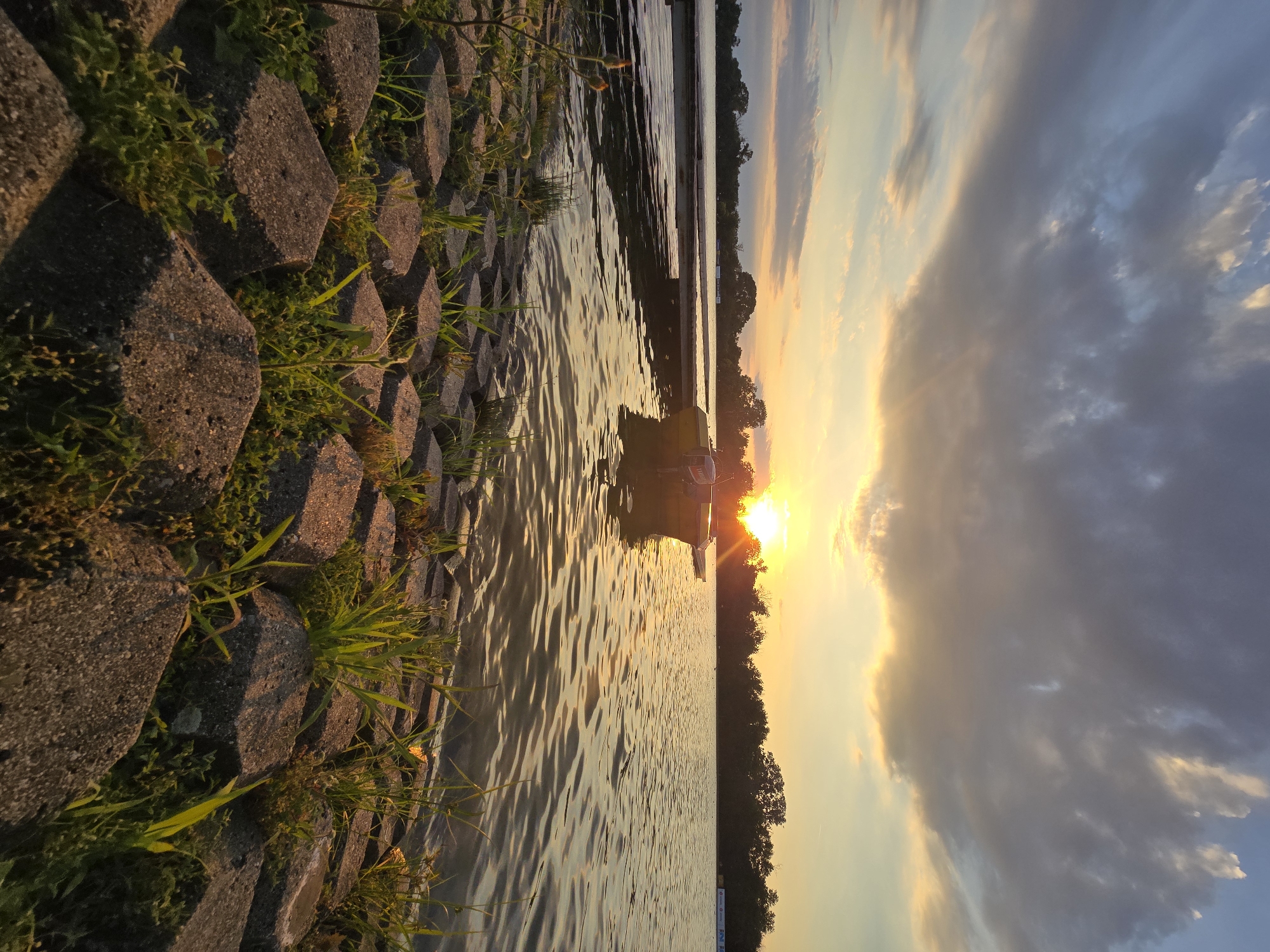 The image depicts a scenic view of a body of water, likely a river or lake, at sunset. The foreground is dominated by a textured, gray, interlocking stone embankment, with patches of vibrant green grass growing between the stones, suggesting a waterfront park or a landscaped riverbank.
The water's surface reflects the warm hues of the setting sun, creating shimmering patterns of light and color. A narrow, dark strip of trees forms a natural horizon line across the middle distance. Above the trees, the sky is a dramatic canvas of swirling clouds, transitioning from a bright, golden-orange glow near the horizon to deeper blues and grays as it ascends. The sun itself is a brilliant, concentrated orb, casting a warm light that illuminates the clouds and the water.
In the water, a small, white boat or raft is visible, with a person silhouetted against the bright light of the setting sun. The presence of the boat and the tranquil atmosphere suggest a peaceful moment, possibly recreational. The overall mood is serene and contemplative, with the vibrant colors of the sunset evoking a sense of beauty and the end of the day. The location context of Plovdiv, Bulgaria, does not provide specific visual cues within the image itself. There is no visible text in the image.