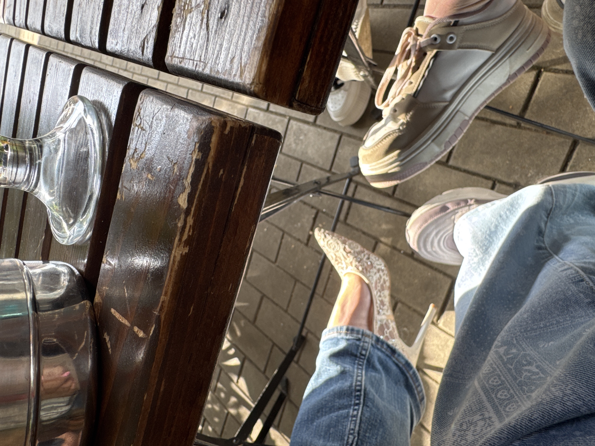 The image presents a close-up, angled view of an outdoor setting in Unknown, Unknown, featuring elements of street furniture and personal items, seemingly captured from an elevated perspective. A wooden slatted table occupies a significant portion of the left side, bearing the base of a glass object and a metal container or item. Below the table, the scene transitions to a paved surface, perhaps a sidewalk or patio.
Two pairs of footwear are distinctly visible. One pair is lace, pointed-toe high heels, worn with a blue denim jean leg. The second is a pair of sneakers, also accompanied by denim. The feet are positioned as if the individuals are seated at the table, their legs casually placed. The background texture and lighting suggest an outdoor setting under natural light.