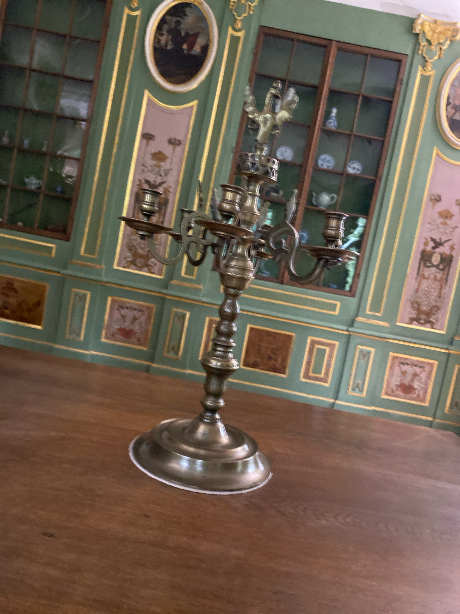 The image shows a dimly lit, ornate room, likely a museum or historic building. In the foreground, a brass candelabra stands on a dark wooden table. The candelabra has an intricate design with multiple arms and candle holders, topped with a bird-like sculpture. The background features a wall with panels of green and pink, decorated with gold trim. There are large, mullioned windows that reflect light, and on the wall, several framed oval paintings and decorative panels are visible. The overall impression is one of historical grandeur and opulence. This room is located in the Hermitage Museum in Saint Petersburg, Russia.