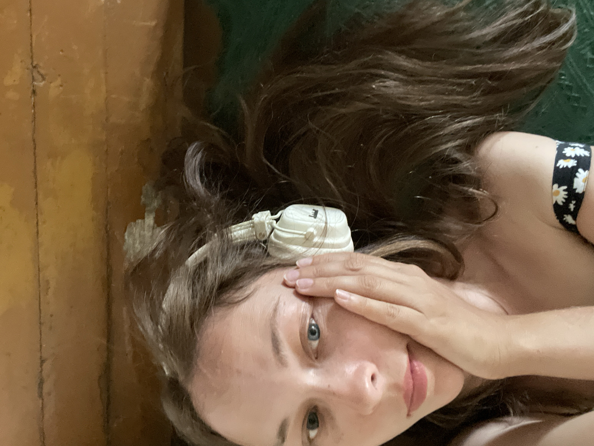 The image shows a close-up, overhead shot of a young woman lying down with her head on a green surface, possibly a carpet or fabric. Her long, dark brown hair is spread out around her head. She is wearing white headphones with a white cord that runs across her forehead. Her right hand is covering her right eye, and her left eye is visible, appearing to be blue. Her skin is fair, and she has some freckles on her cheeks and nose. Her lips are slightly parted. She is wearing a black top with a white floral pattern. The background on the left appears to be a wooden surface, possibly a floor or wall, with a yellowish-brown hue and visible seams. The overall lighting suggests an indoor setting, possibly during the day given the brightness of her skin. The mood appears relaxed or pensive. No visible text is present.