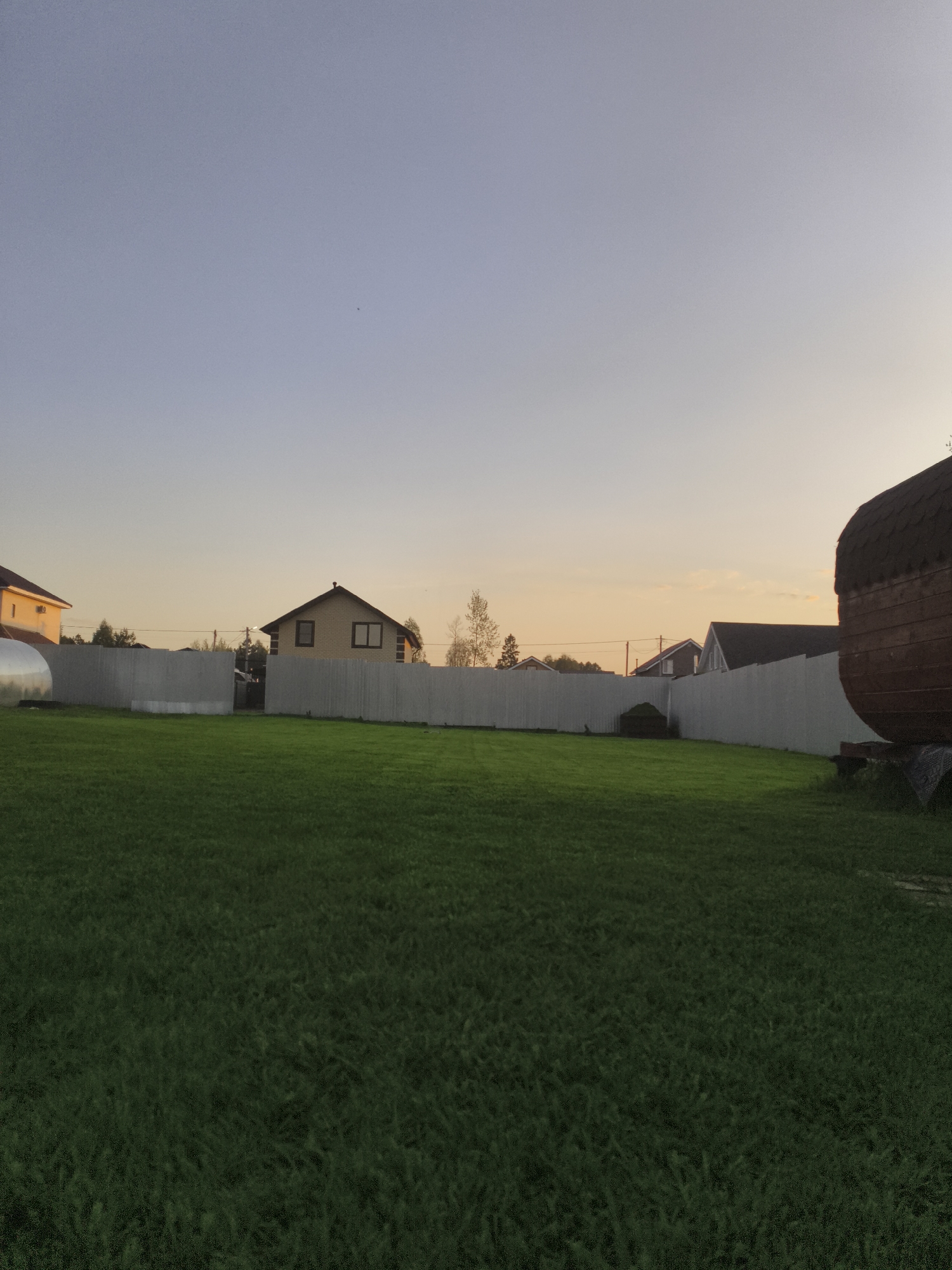 The image depicts a residential area in Русский Вожой, Russia. The foreground consists of a large, well-maintained lawn of green grass. A white fence runs across the midground, separating the lawn from neighboring properties. Several houses are visible behind the fence, constructed in varying architectural styles. In the right side of the image, there is a sauna. The sky transitions from blue to a yellowish-orange hue, indicating either sunrise or sunset. No people are visible in the scene.