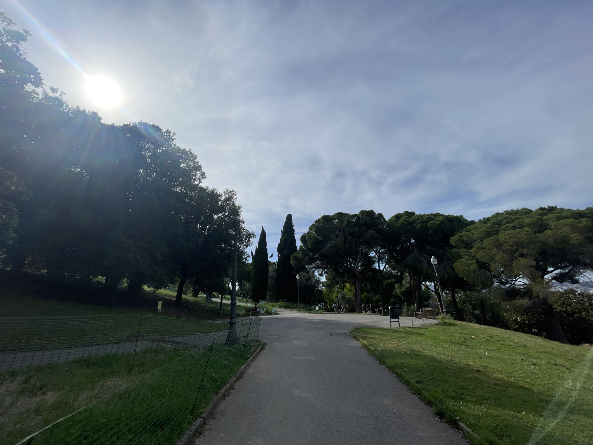 This image depicts a sunlit outdoor scene within a public park, characteristic of Montjuïc in Barcelona, Spain, under a partially cloudy sky. A dark grey asphalt path, slightly worn, extends from the foreground towards the upper center, branching slightly into the mid-distance. To the immediate left of the path, a green-coated wire mesh fence separates it from a downward sloping, deeply shaded grassy area with mature trees; a faint yellow-green marker is visible within this shaded section. To the right of the path, a gently ascending grassy slope features scattered patches of brighter green grass. The top-left quadrant of the sky is dominated by a bright sun, emitting strong lens flare and visible sunbeams, partially obscured by dense, dark green broadleaf trees along the left edge. The sky transitions to a lighter blue-grey with thin, wispy clouds towards the right. Tall, slender cypress trees are visible in the mid-ground, flanked by other mature evergreen trees with full canopies. Along the path, several dark lampposts are present, one on the left with a decorative base, and another further ahead on the right. A dark bench is situated on the right side of the path, and further back on the left, one individual wearing light-colored clothing is seated on another bench beneath a tree. A dark public trash receptacle is positioned on the right side of the path beyond the first bench.