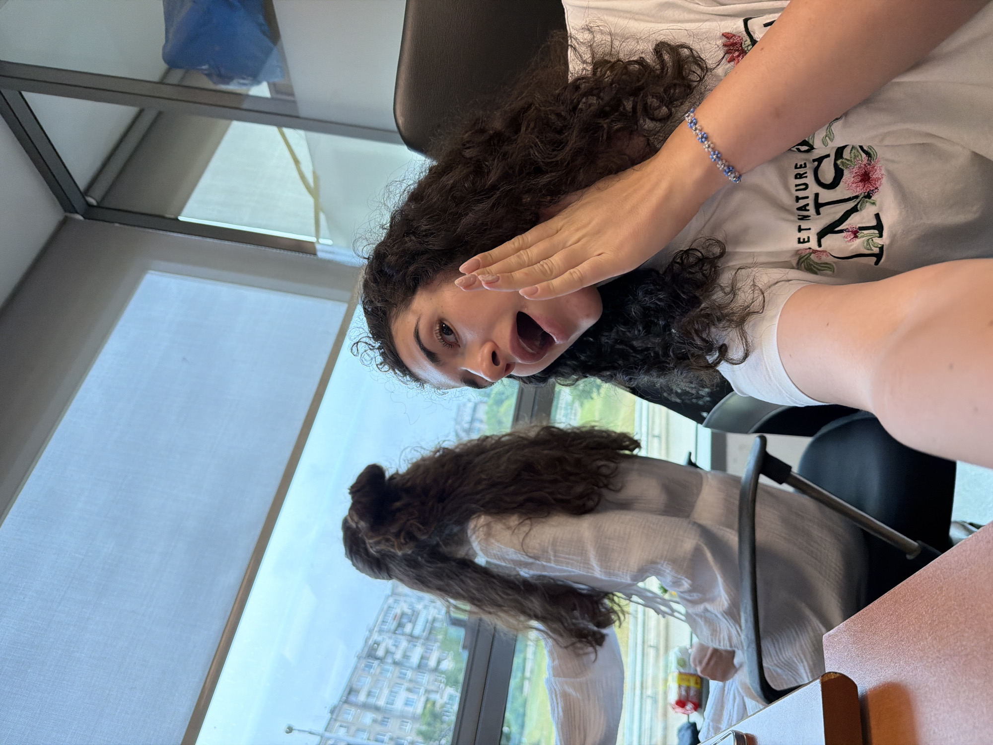 The image depicts two young women with dark, curly hair in what appears to be an office or study environment, with large windows overlooking an urban landscape. The woman in the foreground, on the right, is looking directly at the camera and has her right hand raised to her cheek, with her mouth open in an expression of surprise or excitement. She is wearing a white t-shirt with floral embroidery and the text "ETNATURE INS". A beaded blue bracelet is visible on her left wrist. The woman in the background, on the left, is facing away from the camera, looking out the window. She is wearing a loose-fitting white shirt and her hair is styled in a half-updo. The window offers a view of a cloudy sky and buildings, suggesting an urban setting. Based on the visual cues and the text on the t-shirt, the location is identified as Sofia, Bulgaria. The desk in the foreground is made of wood, and office chairs are visible.