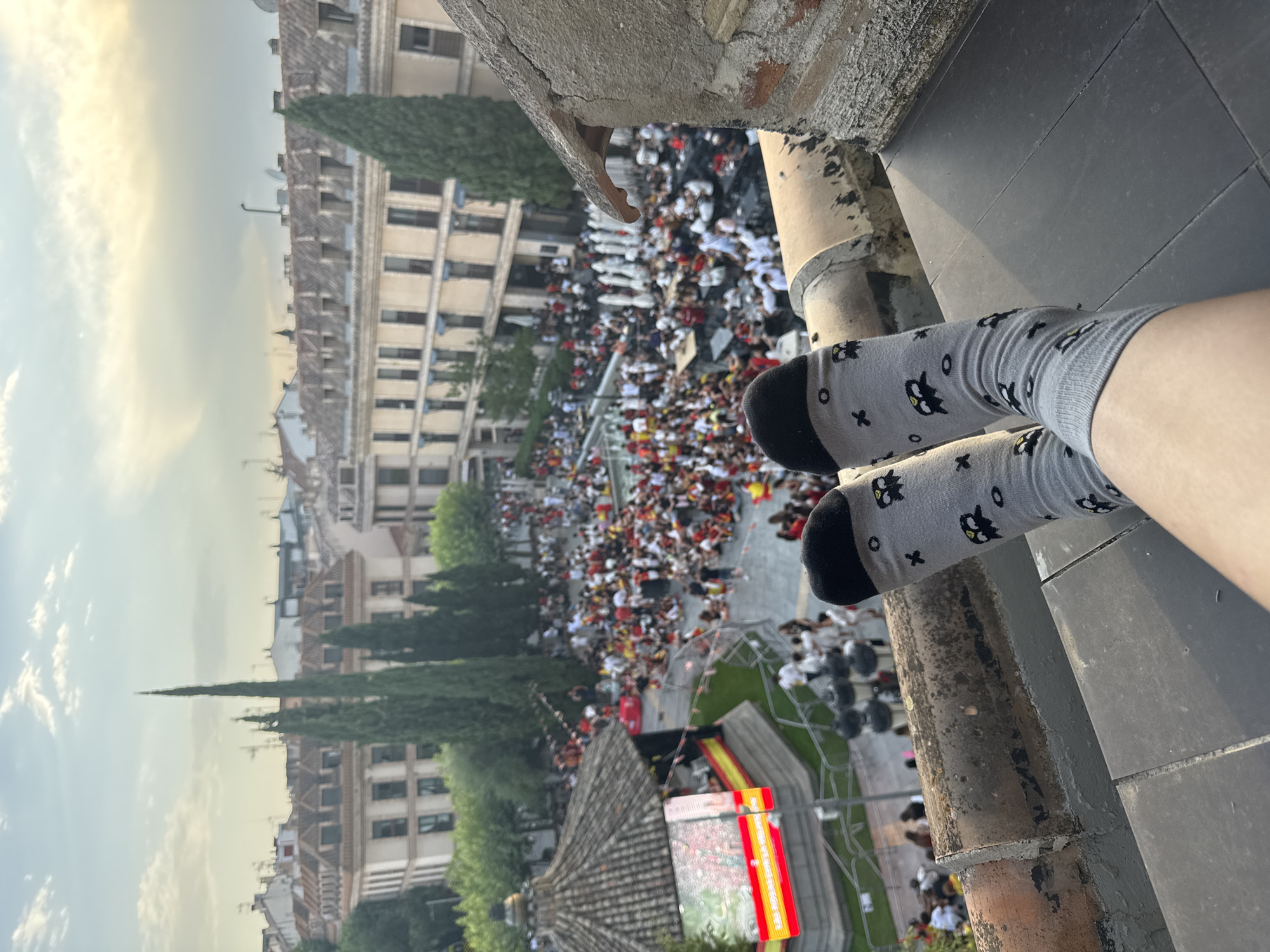 A first-person perspective shot shows a pair of feet clad in gray socks with a pattern of cartoon owls and crosses, resting on a dark, textured surface. The socks are worn by an individual positioned on a rooftop or elevated vantage point. In the background, a bustling city square is visible. Buildings with tiled roofs and multiple windows line the square. A crowd of people, many wearing red and yellow, are gathered in the square, some seated at tables. A stage or performance area is set up on the left side of the square, adorned with Spanish flags and featuring a large screen displaying what appears to be a map. Cypress trees are interspersed among the buildings, and the sky above is partly cloudy with the sun low on the horizon. The scene depicts a public gathering or event taking place in an urban environment, possibly a city square in Las Rozas de Madrid, Spain.