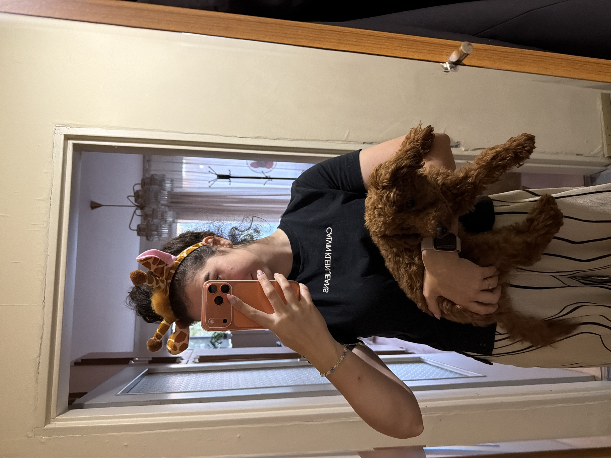 A young woman with curly brown hair, wearing a black t-shirt with white lettering, a beige and white striped skirt, and a giraffe-ear headband, holds a small, fluffy brown dog in her left arm. She is taking a selfie in a mirror. The background shows an interior room with a door on the left and a wardrobe on the right. A chandelier is visible in the reflection of the mirror. This appears to be a residential setting.