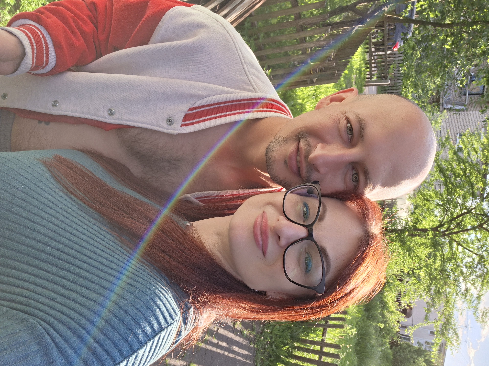 This is a close-up selfie of a man and a woman in an outdoor, rural setting. The man, who is bald with light skin and a goatee, is on the left. He is wearing a white and red varsity-style jacket, partially unbuttoned, revealing his chest. The woman, on the right, has long, reddish-brown hair and is wearing glasses and a teal ribbed shirt. A rainbow-like light flare crosses both of them. Behind them, a wooden fence is visible, along with trees and a building in the distance. The overall environment suggests a village or countryside, consistent with the location in Забалоцкі сельскі Савет, Belarus. The lighting indicates it is daytime, likely sunny, as suggested by the strong shadows and the light flare.