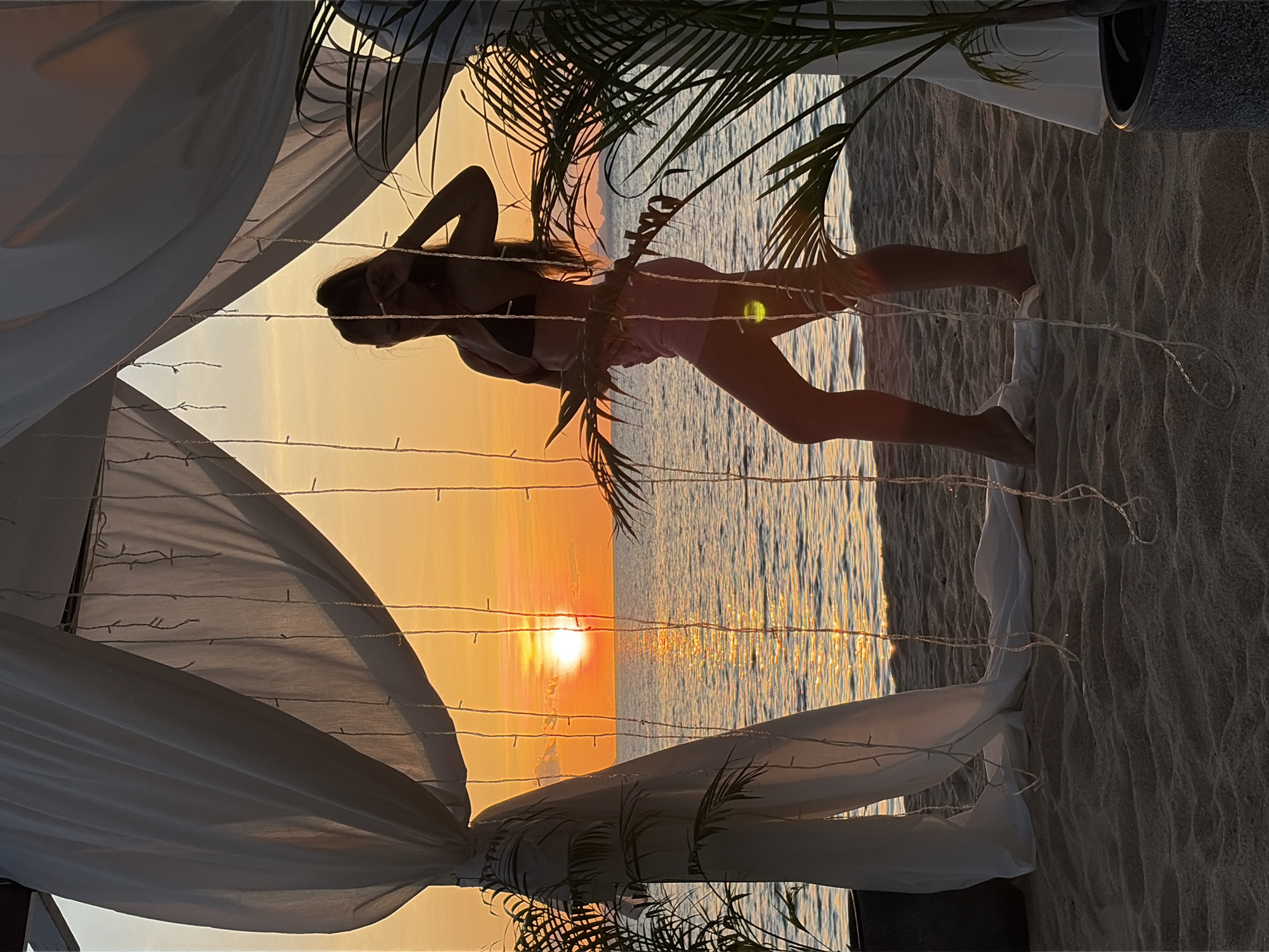 A silhouette of a woman stands on a sandy beach in Baan Taling Ngam, Thailand, at sunset. She is wearing a bikini top and pink shorts. The woman is partially framed by white draped fabric and strings of lights, suggesting a cabana or decorative setup. Palm fronds are visible on either side of the frame, with one plant in a dark pot on the left and another on the right. The sun is setting over the ocean, casting a warm glow and reflecting on the water. The sky is a vibrant orange and yellow. The sand is textured with ripples.