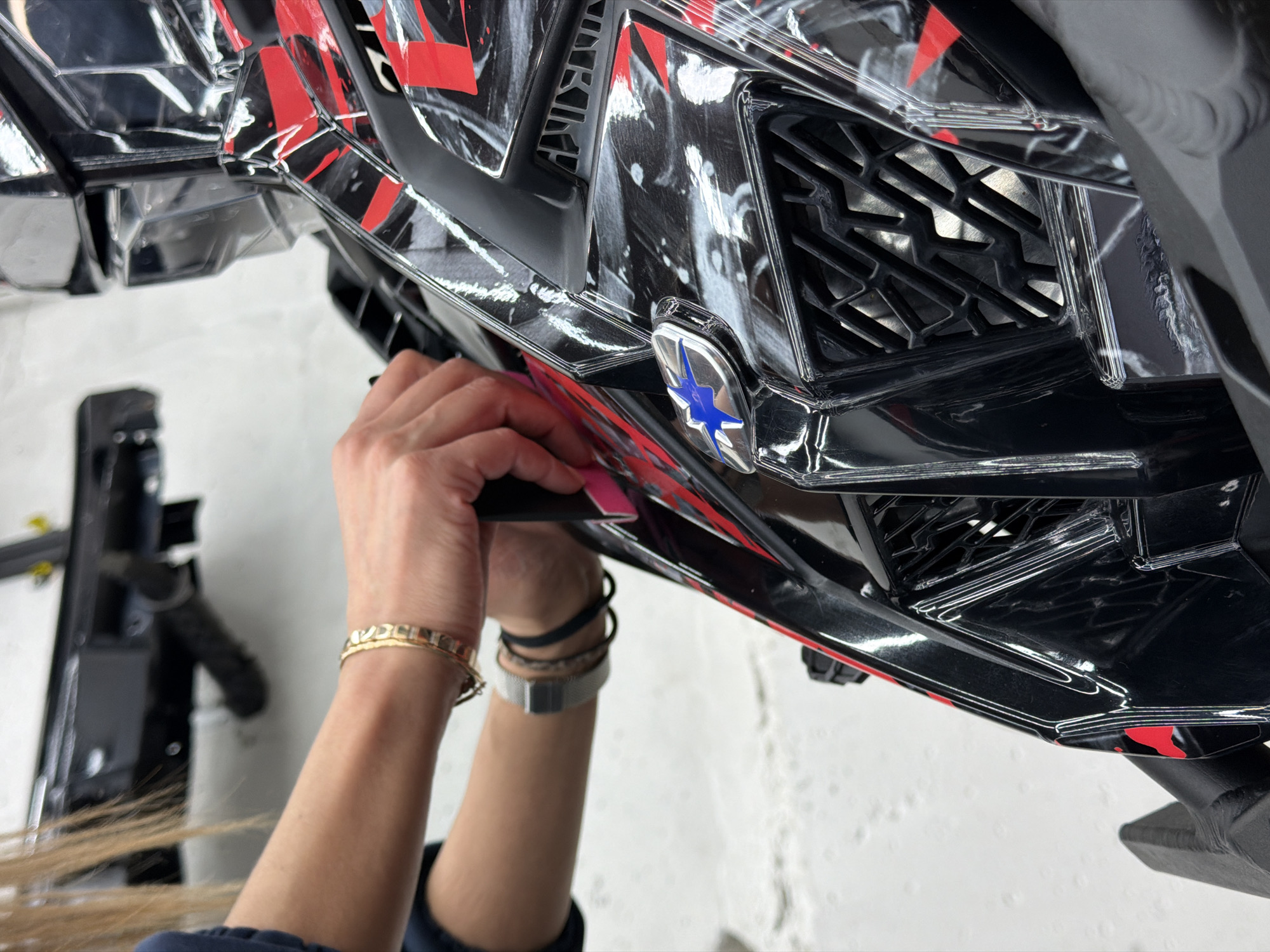 A close-up shows a person's hands applying something to the underside of a black vehicle part with red and white markings. The part features a grid-like design. A silver emblem with a blue star-like shape is visible. The person has bracelets and a watch on their left wrist. A partially visible black object rests on the left. The background is a white, clean surface. There is no explicit information in the image to confirm the location is Krasnoyarsk, Russia.
