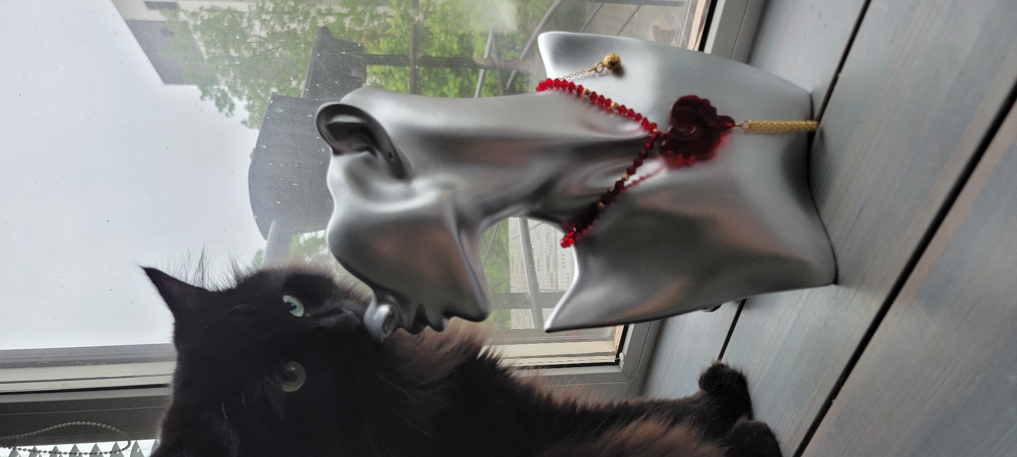 A black cat is seen looking up at a mannequin head positioned on a windowsill. The mannequin is silver and adorned with a red beaded necklace featuring a rose detail. The window reveals an outdoor scene with trees and a building. The window frame is white, and the windowsill is gray with a wood-grain texture. The scene is well-lit, possibly by natural light from the window.