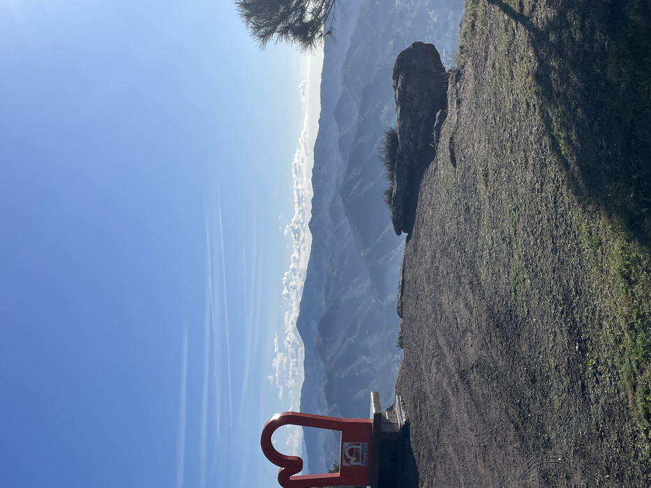 The image shows a view from a clifftop near the town of Álora, Spain. The location is a rocky outcrop with a red metal sculpture shaped like a large, stylized "G" at the bottom of the image. The sculpture is a symbol of the town, called "El Gran G." The background shows a blue sky with wispy clouds, and a distant view of rolling hills and mountains. The image is taken from a slightly oblique angle, looking downwards and out toward the mountains.