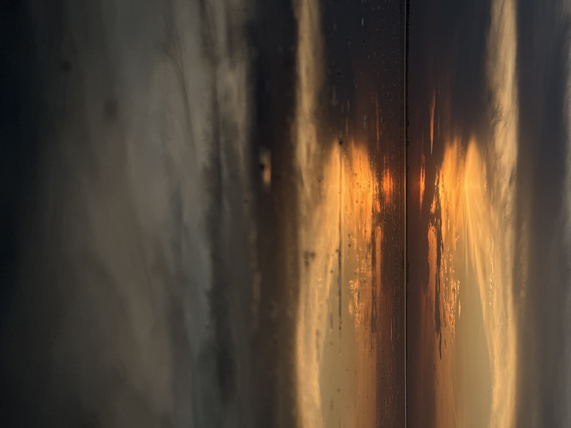 The image shows a reflection of a sunset with clouds in a window or a shiny surface. The reflection is horizontally flipped. The sunset features shades of orange and yellow. The background is darker, with hints of gray and white clouds. The scene seems to be captured in two panes due to the vertical division, suggesting that the image may be a view of the reflection from a window. The image does not contain any identifiable people, subjects, or actions. The location is not identifiable based solely on the visual information provided.