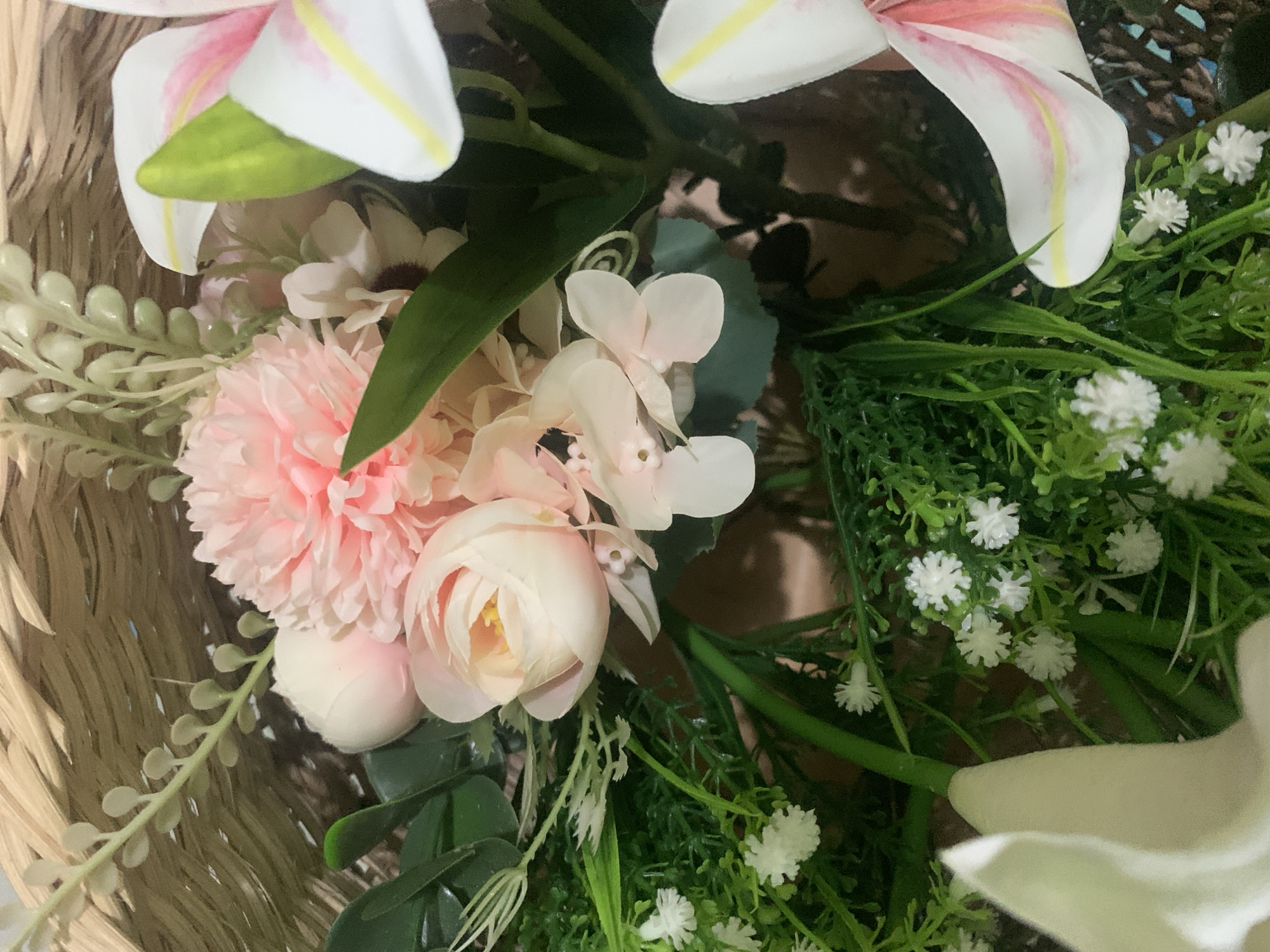 A close-up shot shows a floral arrangement in a woven basket. The arrangement features various artificial flowers, including pink carnations, delicate white blossoms resembling baby's breath, and white lilies with pink and yellow accents. Lush green foliage and some lighter green decorative stems fill out the composition. The basket appears to be made of natural fibers and has a visible weave pattern. The background is dark and out of focus, suggesting an indoor setting. No people or specific landmarks of Kuala Pembuang, Indonesia are visible in this image.
