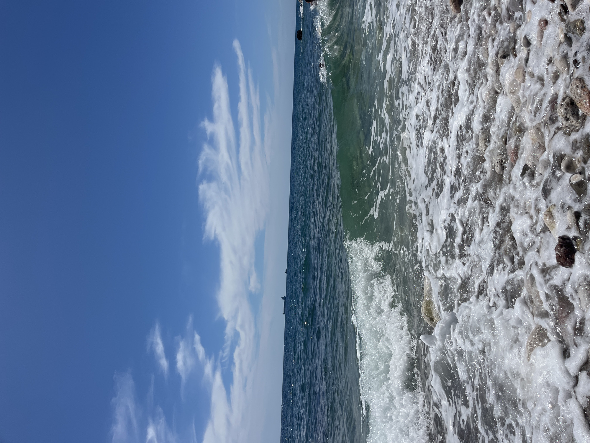 A wide shot captures a tranquil sea under a clear blue sky with wispy white clouds, suggesting a pleasant day. The water is a deep, rich blue, with visible waves breaking near the shore. The foreground is a pebbly beach, partially covered by foamy sea spray as a wave recedes. In the distance, three cargo ships are visible on the horizon, indicating maritime activity. The scene is likely located on a beach in Bar, Montenegro, known for its scenic coastline and clear waters. No people are clearly visible, but the presence of ships suggests the area is frequented.