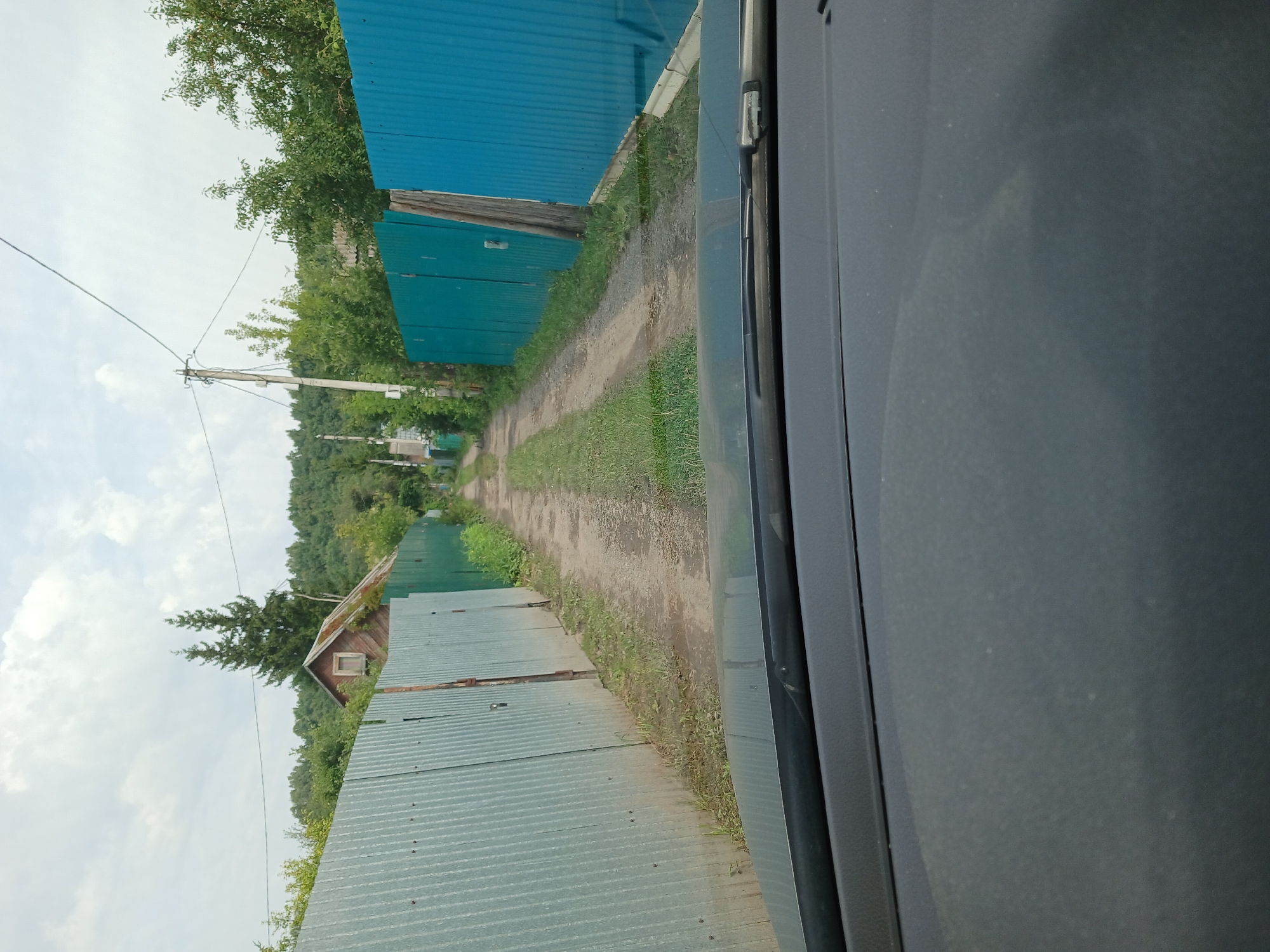 The image shows a dirt road in a village, possibly in Vasilyevo, Russia. The road is flanked by corrugated metal fences on both sides. The fence on the left is silver-gray, and the fence on the right is bright blue. There are some trees and vegetation growing alongside the road and behind the fences. In the distance, a small, weathered wooden house with a mossy roof is visible behind the left fence, with two tall evergreen trees behind it. A utility pole with wires is also present. The view appears to be from inside a vehicle, looking forward through the windshield. The sky is overcast with clouds.