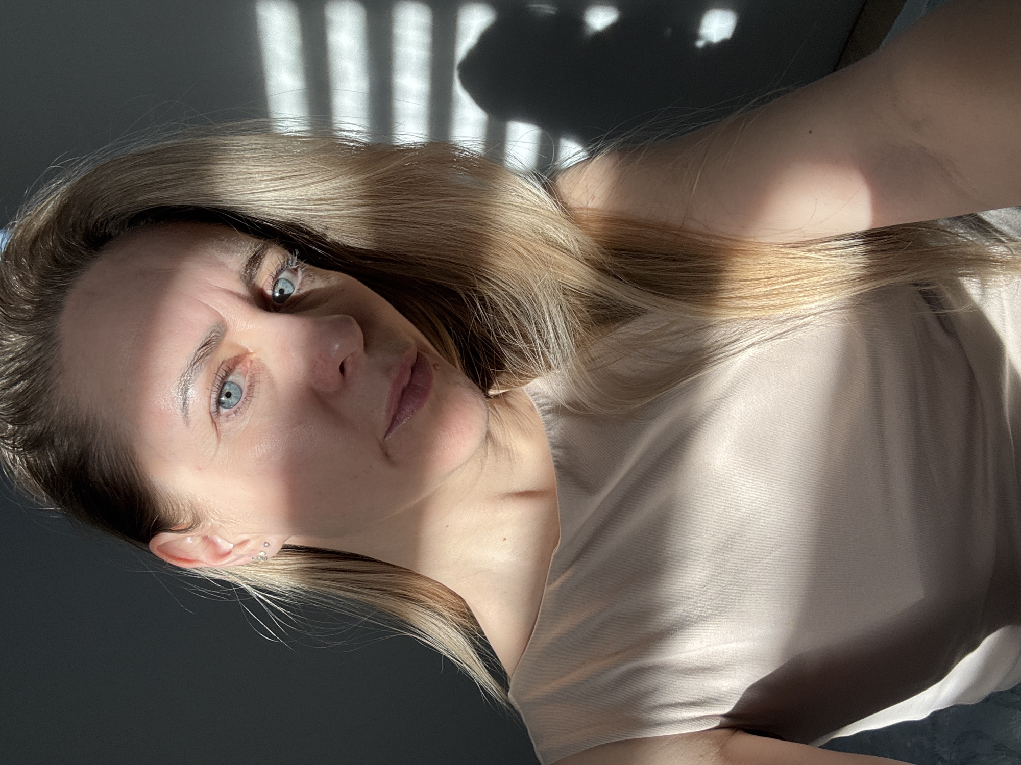 The image is an indoor, self-portrait of a woman with blonde hair and blue eyes, captured in Chaikovsky, Russia. She is looking directly at the camera, her head tilted to the side, wearing a light-colored, possibly beige or pale pink, t-shirt or top.
Bright sunlight streams through a window, casting distinct striped shadows onto a dark wall in the background and across her hair, face, and clothing. This strong interplay of light and shadow highlights her features and hair, suggesting it is either morning or late afternoon. The visible shadows of window blinds or slats indicate a sunny day outdoors.
The woman's expression appears neutral or slightly serious, with a direct and steady gaze. Notable details include fine lines around her eyes and forehead, and small, multiple piercings in her left ear. A small mole is visible on her neck. Her blonde hair is illuminated by the sunlight, creating bright highlights. No visible text is present in the image.