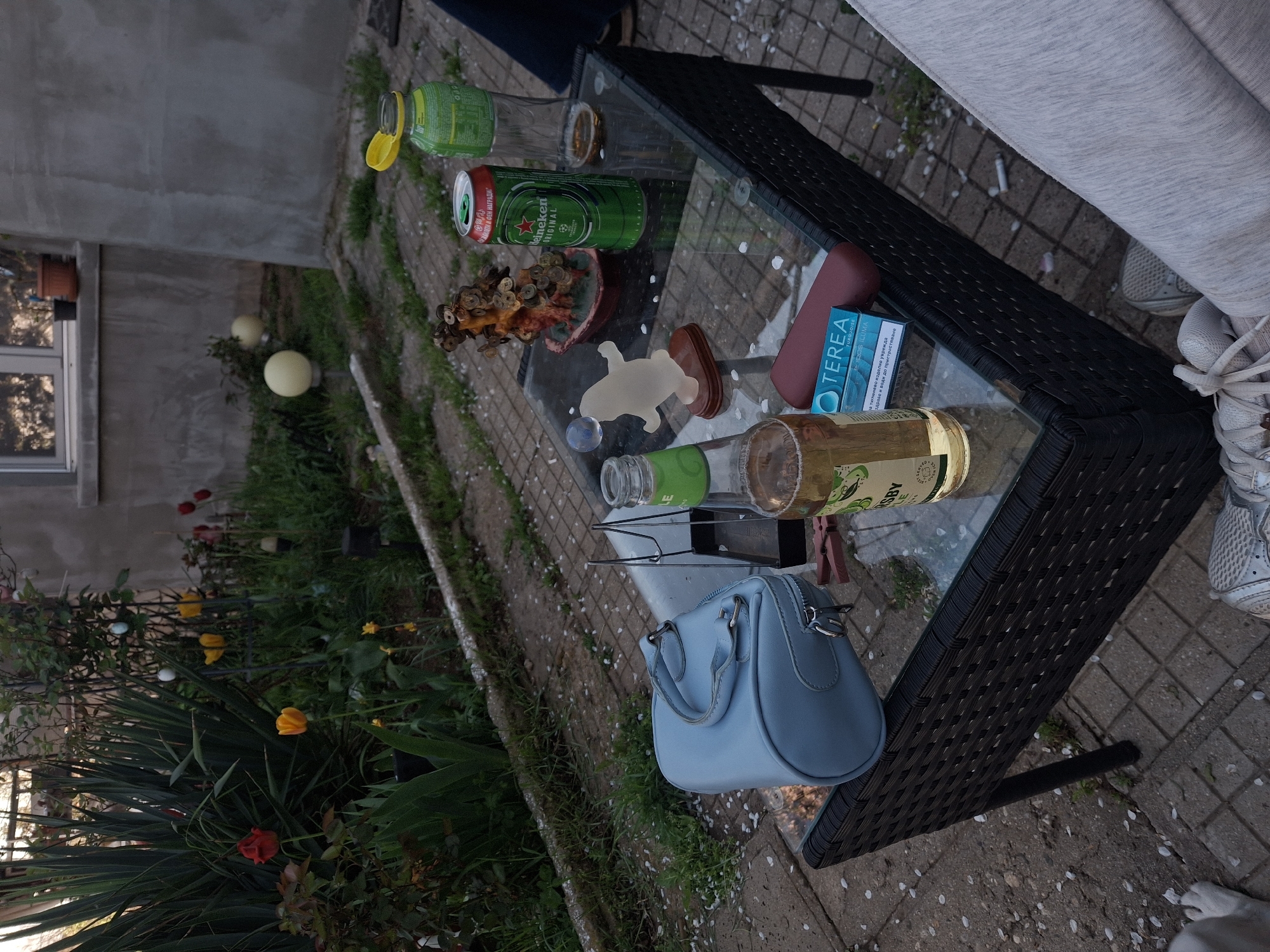 A medium shot depicts an outdoor setting with a glass-topped table holding various items. A Heineken can, a decorative coral sculpture, and an object shaped like a turtle are positioned towards the center of the table. A pack of "TEREA TURQUOISE" cigarette cartridges and two glass bottles, one containing a yellow liquid, also rest on the table. A light blue handbag is suspended from the edge of the table. A person wearing grey sweatpants and white sneakers stands adjacent to the table. The table is surrounded by a brick-paved area with scattered white flower petals. Beyond the paved area, lush greenery is visible, including ornamental grasses, flowering bushes with red and yellow flowers, and spherical garden lights. A stone wall and a building with a window can be seen in the background. Given the style of the architecture and landscaping, the location is possibly Buhovo, Bulgaria.