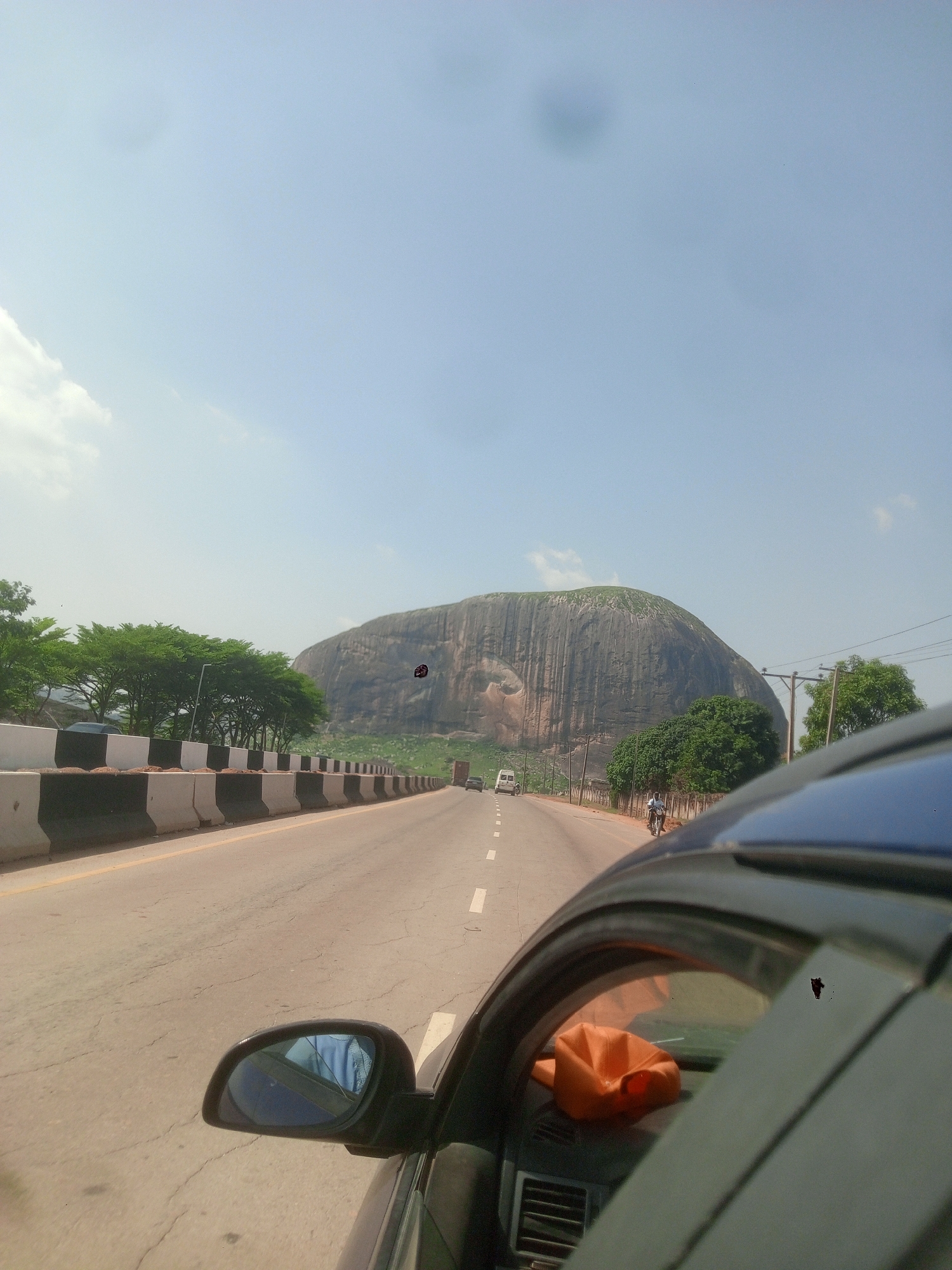 A road with a large rock formation in the background is visible. The road is paved and has a barrier on the left side. A white van is driving on the road towards the rock formation. There is a man on a motorcycle on the right side of the road. A car is partially visible in the foreground. The sky is blue with some clouds. The location is Unknown, Unknown.