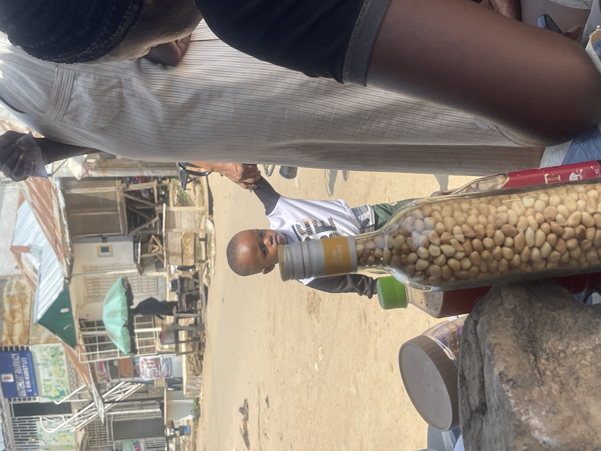 In a sun-drenched street scene in Abuja, Nigeria, a young child, appearing to be between three and four years old, stands holding the hand of a taller person, possibly an adult, dressed in a flowing, light-colored garment. The child is wearing a white t-shirt with black and gold striped accents and green shorts. They are positioned outdoors on a dusty, unpaved surface, with various elements of a local marketplace visible in the background. These include a green umbrella providing shade, market stalls with visible goods, and buildings with corrugated metal roofs. In the foreground, a large glass bottle filled with what appears to be peanuts or similar legumes stands prominently, along with other bottles and containers, suggesting a retail or street vending environment. The overall impression is a snapshot of daily life in a bustling, possibly informal, commercial area.