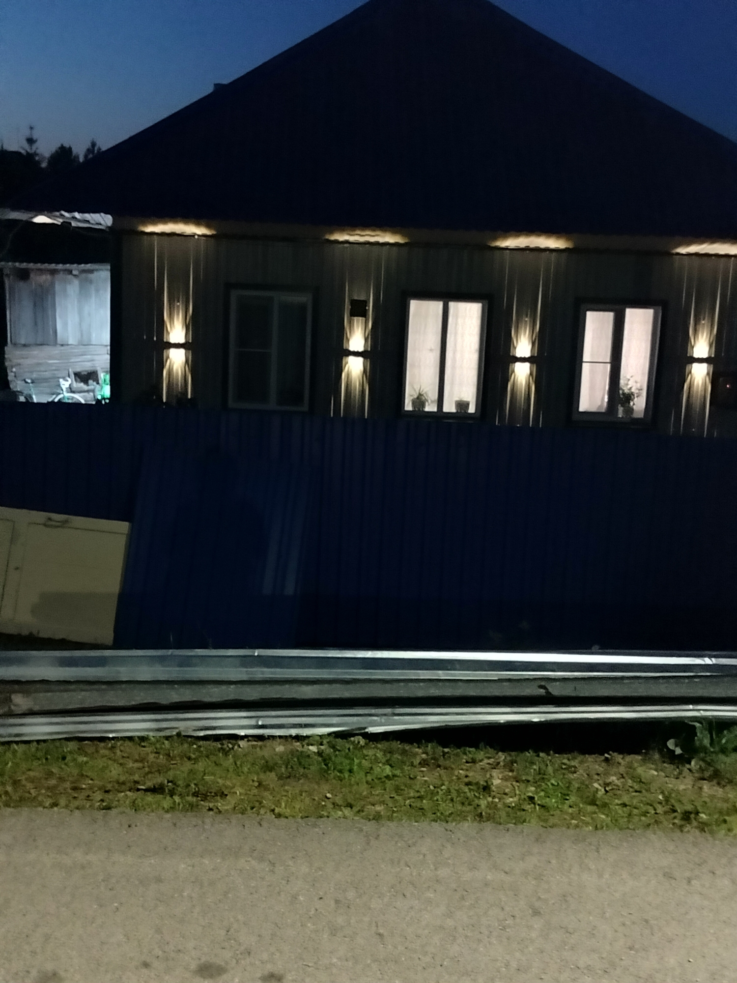 The image depicts the exterior of a two-story building at dusk or night, illuminated by outdoor lighting. The building has a dark-colored roof and a facade covered in vertical metal siding, likely corrugated. Three rectangular windows are visible on the upper level, each with translucent curtains, suggesting they are indoors. Above each window, and between them, are decorative outdoor lights that cast a warm glow onto the facade. The building is partially obscured by a tall, dark blue corrugated metal fence in the foreground. To the left of the main building, a section of a less finished structure, possibly a shed or a garage, is visible. This structure also has a corrugated metal exterior.

In front of the fence, a light-colored, rectangular object, possibly a construction panel or a door, is leaning against the fence. Beyond the fence and the building, a dark sky with a hint of twilight is visible, with a few dark tree silhouettes at the far left edge. The foreground consists of a strip of green grass and a paved surface, likely a road or sidewalk. A metallic barrier or railing is situated between the grass and the paved area. The overall scene suggests a residential or semi-commercial area on the outskirts of Yashkino, Russia, at a time when artificial lighting is necessary for visibility. No people are visible in the image, and no specific actions are depicted.
