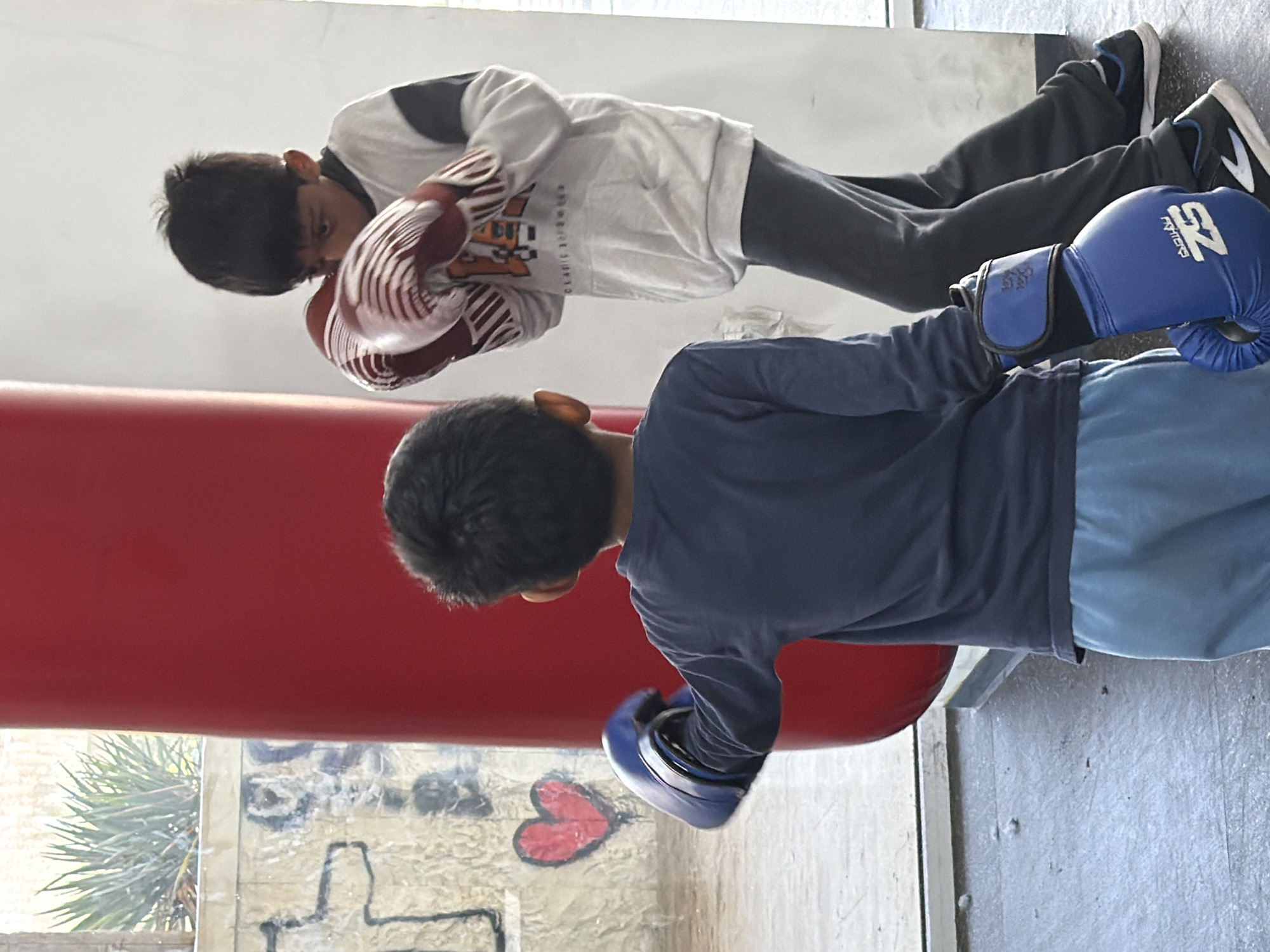 The image shows two young boys boxing. The boy in the foreground is wearing a navy-blue long-sleeve shirt and light-blue shorts. He has blue boxing gloves and is punching a red punching bag. The boy in the background is wearing a white sweatshirt with a black collar and dark-grey pants. He has red boxing gloves and is also punching the punching bag. The background includes a wall with graffiti and a window. The scene appears to be indoors, possibly in a gym or training facility. The location of the city of Blagoevgrad, Bulgaria is not clear from this image.