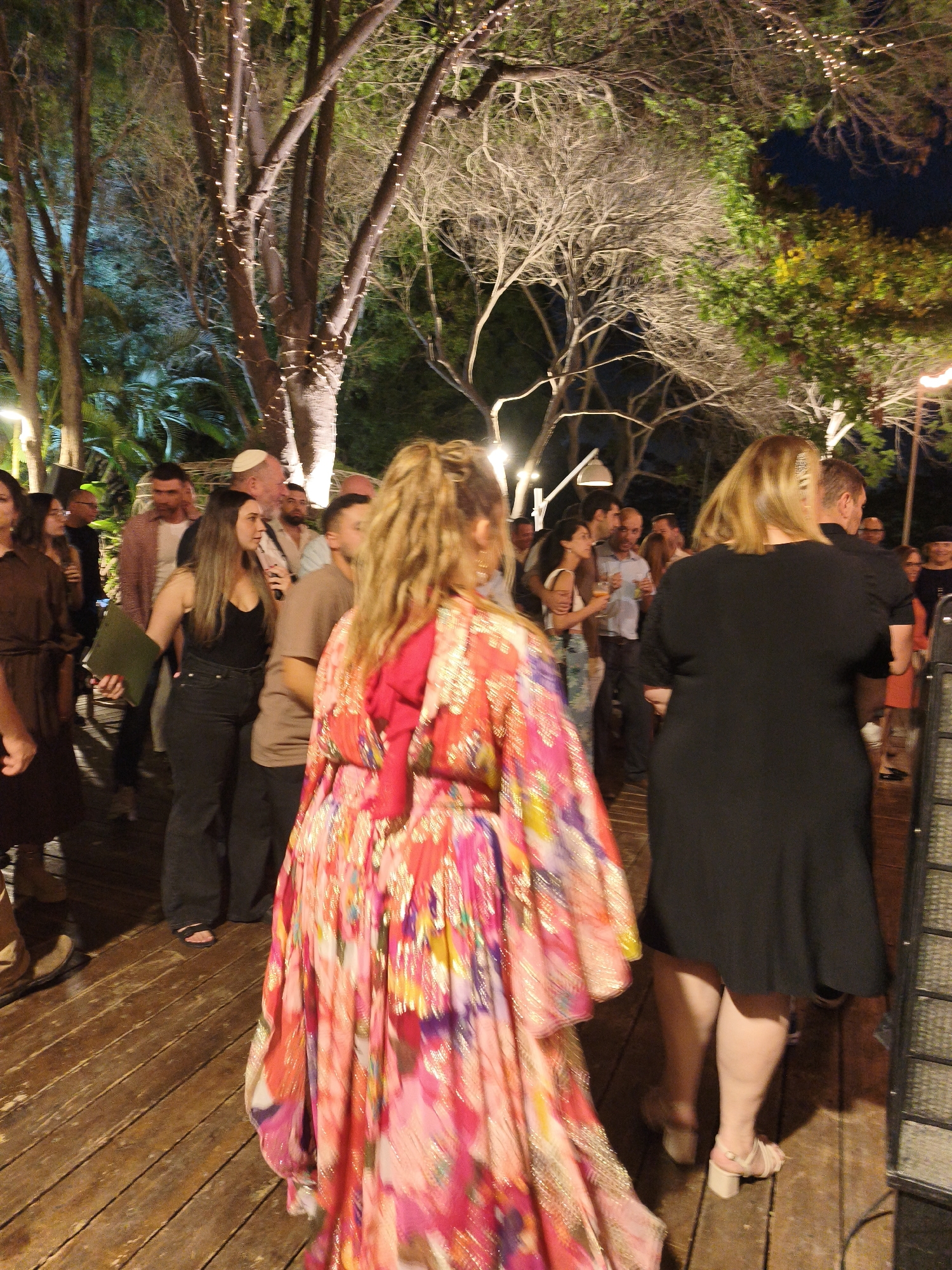 The image depicts an outdoor evening gathering at a venue in Hulda, Israel. In the foreground, a woman wearing a vibrant, floral-patterned dress is seen from behind, her blonde hair styled in a ponytail. To her right, another woman in a black dress is also facing away from the camera, with her legs and feet visible in white heeled sandals.
The background reveals a group of people mingling. Several trees with string lights are visible, suggesting a festive atmosphere. The ground is covered with wooden planks, typical of an outdoor deck or patio. In the distance, more trees and foliage are present, illuminated by the string lights. The lighting conditions suggest it is after sunset, with artificial lights providing illumination. The overall scene appears to be a social event or party.
