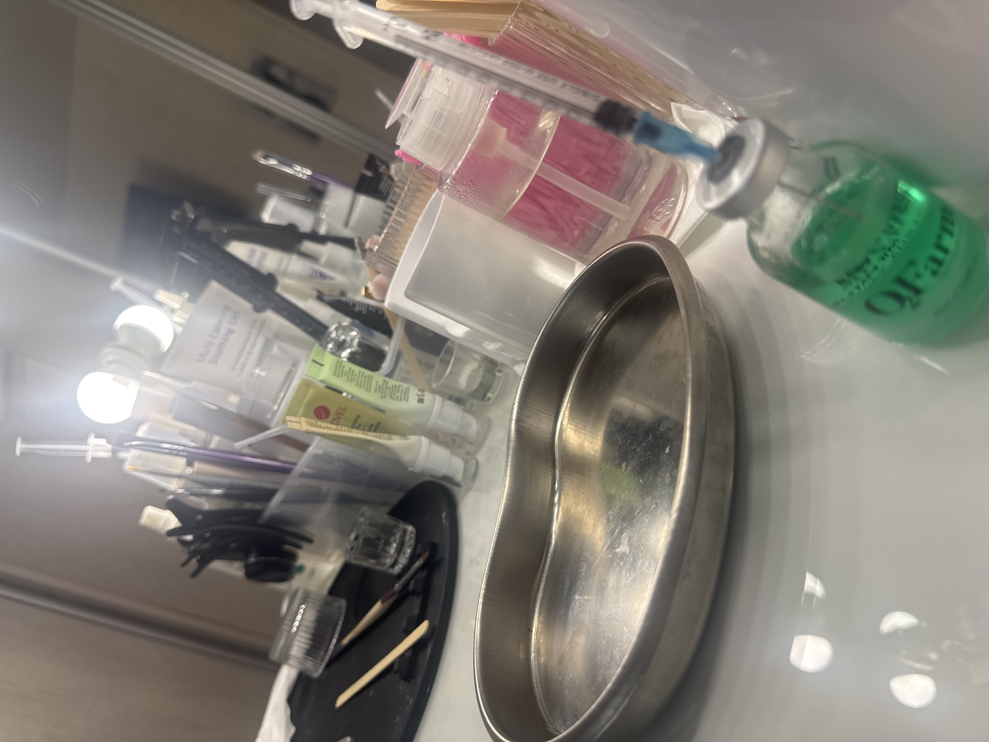 The image depicts a close-up, angled view of a medical or beauty treatment setup. In the foreground, a kidney-shaped metal tray sits on a white surface, reflecting light. To the right of the tray, a small glass vial filled with green liquid is visible, with a syringe inserted into its cap. The vial has text that appears to read "QI Farm." Behind these items, a clear plastic dispenser bottle containing a pink liquid is positioned. Further back, a collection of various beauty or medical tools are arranged in a clear container, including brushes and hair clips. A mirror is visible in the background, reflecting lights and more equipment, creating a blurred and somewhat chaotic impression of a workspace. There are no people visible in the image. The scene does not provide any information that can be used to determine a location in Saratov, Russia.