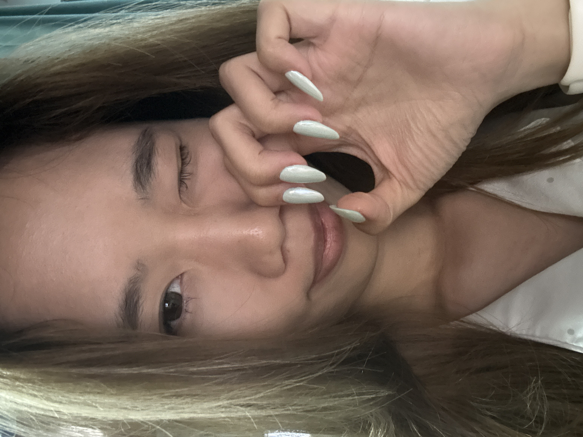 A young woman with tan skin and light brown hair lies horizontally, facing the camera with a slight tilt. Her left eye is partially closed, while her right eye is open and brown. She's making an "OK" sign with her right hand near her face, partially covering her mouth and nose. The nails on her right hand are long, pointed, and painted a light color. She appears to be wearing a white garment. The background is blurred, suggesting a close-up shot. The image does not contain information indicating the location as the Nan'an District of China.