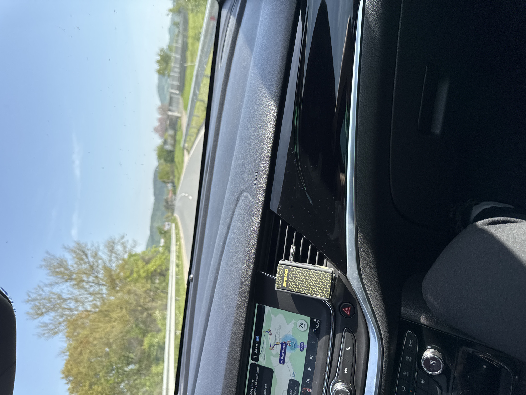The image shows the interior of a car, likely in Daskotna, Bulgaria. The dashboard features a navigation screen displaying a route on ул. Иван Вазов, with a current speed of 76 km/h and a time of 10:29. An air freshener is attached to the air vent. The surrounding environment visible through the windshield includes a road, trees, and clear sky. The image appears to have been taken during a car journey.
