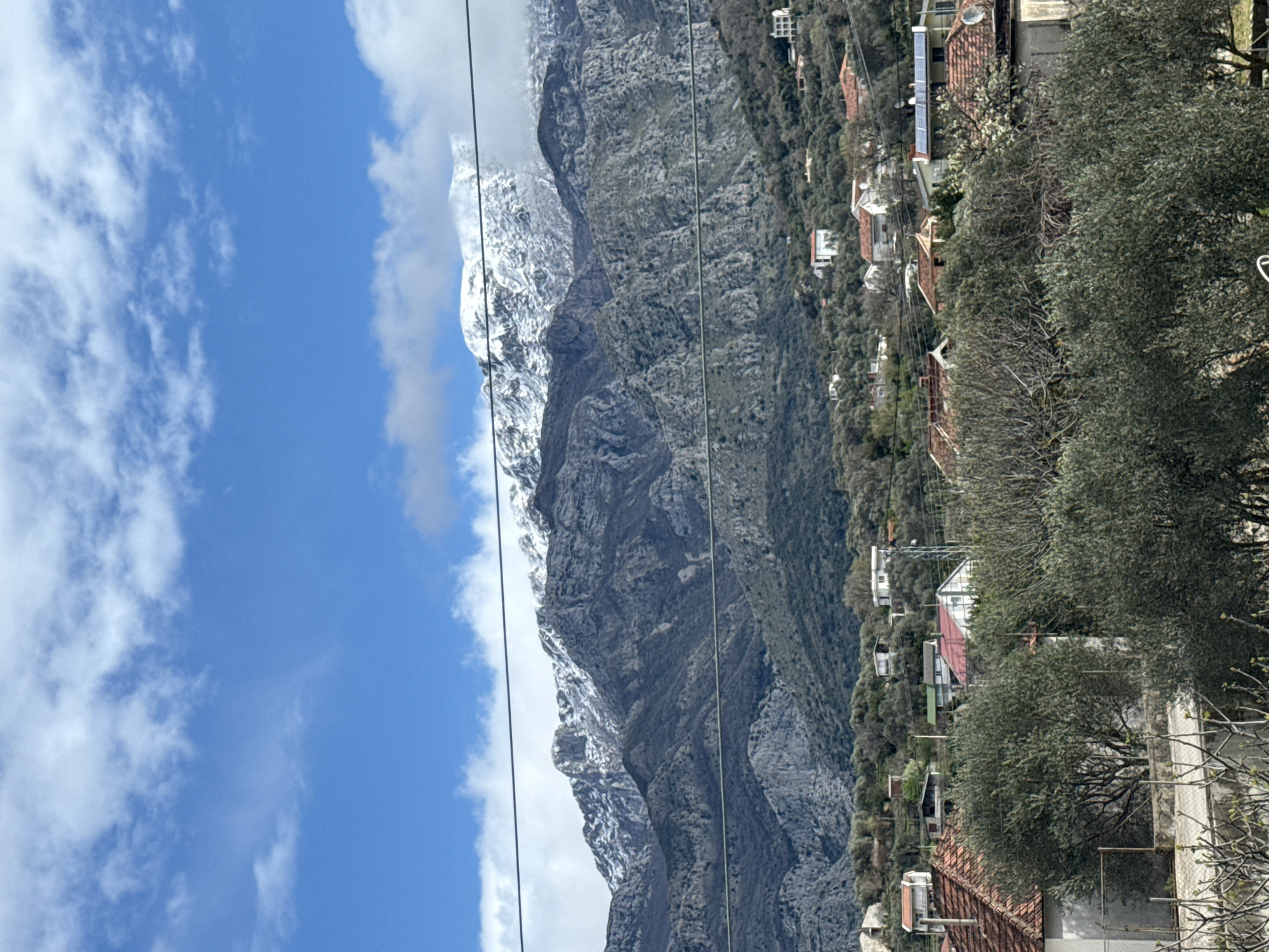This image depicts a mountainous landscape featuring a village under a partly cloudy blue sky. The location is Unknown, Unknown.
In the background, rugged grey mountains rise prominently, with their highest peaks covered in white snow. Some snow-capped areas are partially obscured by white clouds. The mountain slopes below the snowline exhibit exposed rock and sparse, dark green vegetation.
The midground and foreground reveal a residential settlement. Numerous houses with orange or reddish-brown tiled roofs are scattered across the lower mountain slopes and hillsides. These buildings are interspersed among dense vegetation, predominantly olive trees with their characteristic silvery-green leaves, alongside some bare-branched deciduous trees. Utility lines, appearing as dark horizontal wires, traverse the frame in the upper and middle sections. Solar panels are visible on the roof of at least one house in the midground, and a satellite dish is present on another. A chain-link fence is partially visible in the lower-left foreground. No individuals are present, and no active scenes or interactions are depicted. The scene is illuminated by bright daylight.