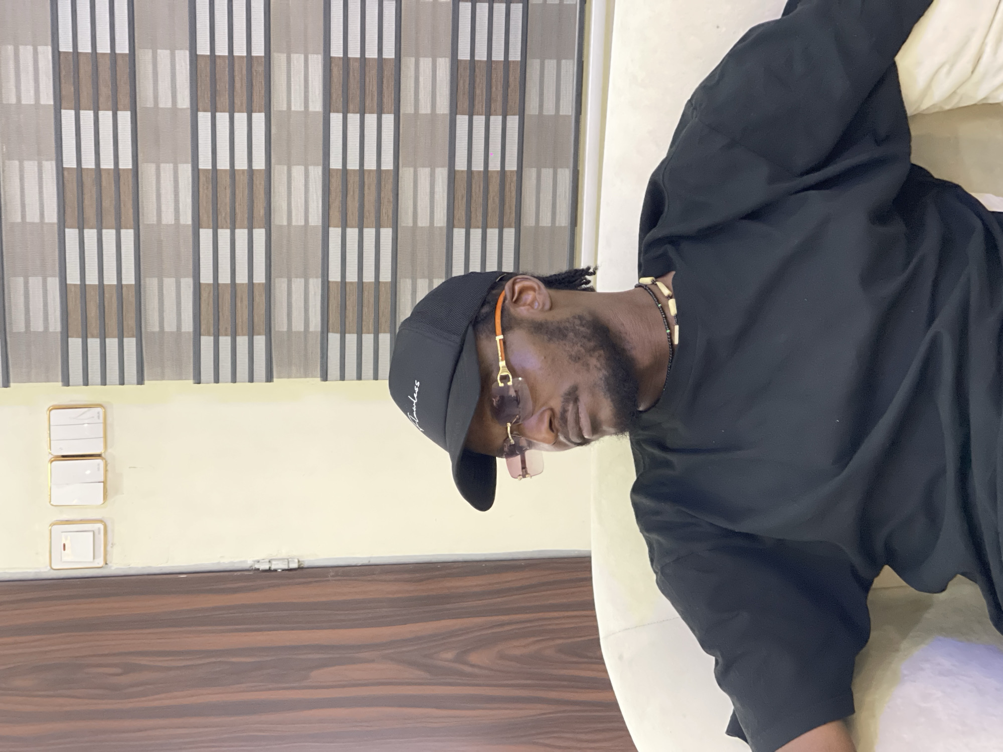 A man is depicted reclining indoors, appearing to be in a relaxed pose. He has dark skin, a beard, and what appears to be dreadlocks or braids partially visible under a black baseball cap. The cap has the word "flawless" written in white cursive script on the front. He is wearing gold-framed sunglasses with reddish-tinted lenses. Around his neck, he wears several beaded necklaces; one is black with small white/cream beads, and another features larger, light-colored wooden or bone beads. He is dressed in a plain black long-sleeved top or sweatshirt.
The man is positioned on a light-colored couch or sofa. In the background, behind him, there is a wall painted in a cream or light yellow hue. On this wall, a set of three white rectangular light switches with gold trim is stacked vertically. The bottom switch has a red indicator light. Above the wall, a set of vertical window blinds with a striped pattern, alternating between light beige/white and darker brown/grey sections, is visible. Below the wall, a surface with a distinct dark brown wood grain pattern, possibly a floor or furniture, is present. The overall setting suggests a comfortable indoor environment, likely a living room or lounge area in Gwarinpa, Nigeria. The natural light visible through the blinds suggests it is daytime.