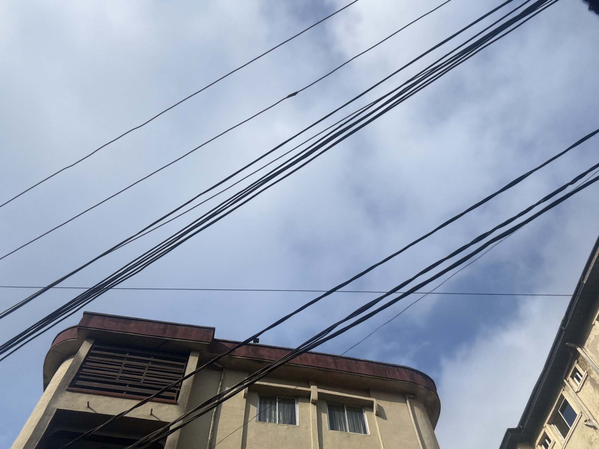 In Onitsha, Nigeria, the image captures a low-angle view of urban infrastructure against a sky with scattered clouds. Prominently featured is the upper section of a light-tan building with red trim, showing two windows with white frames and blinds. A large, dark-slatted ventilation grate is visible near the top of this building. Below it, another building's roofline, also light-tan, is partially visible with windows. A complex network of black power lines stretches diagonally across the frame, some appearing taut and others with slight slack. The overall scene depicts a typical urban environment in Onitsha, characterized by buildings and overhead electrical infrastructure under a cloudy sky.