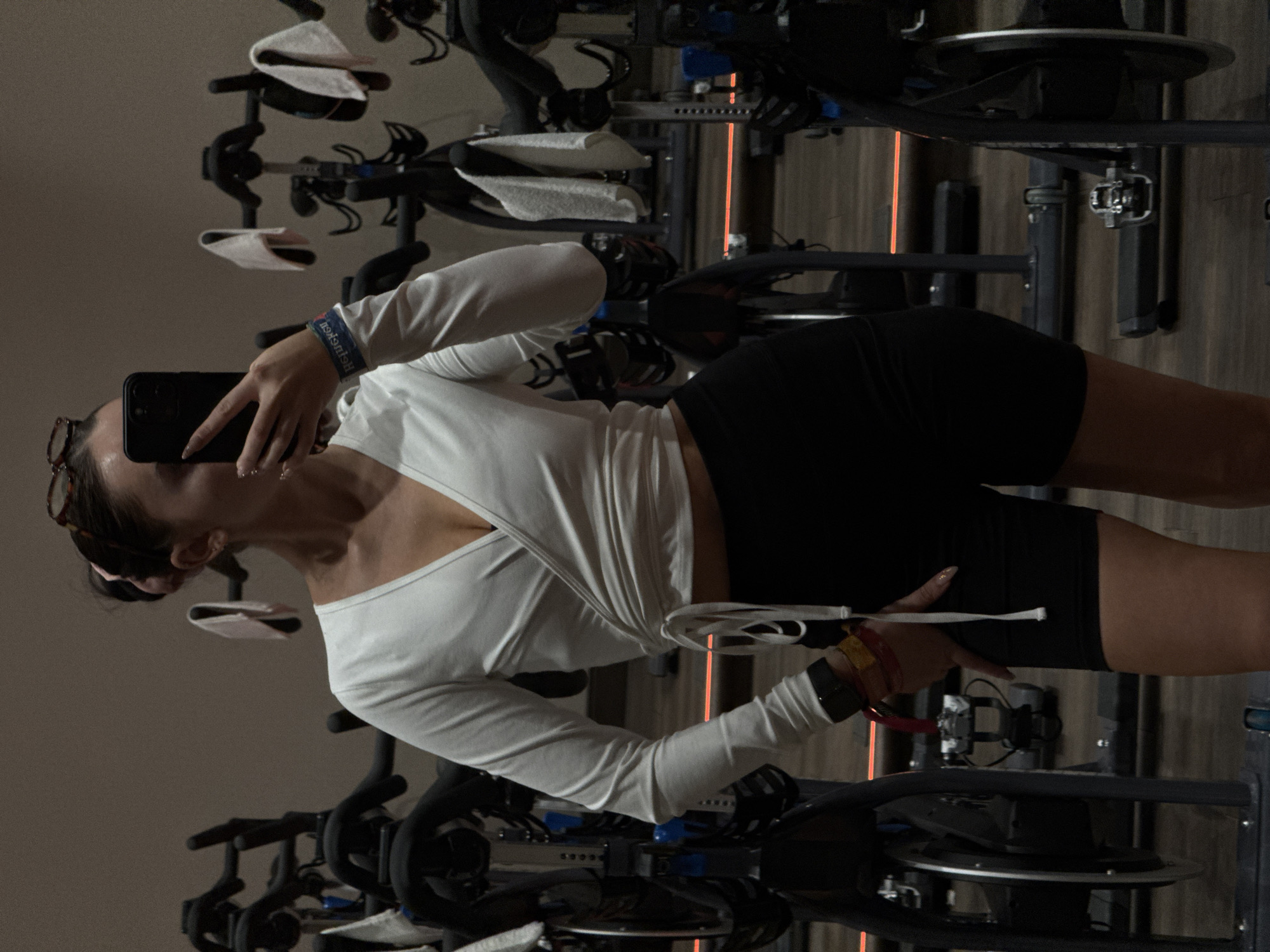 A woman in black exercise shorts and a long-sleeved white wrap top takes a selfie on a stationary bike in a spin class setting. The photo, possibly taken indoors, shows multiple stationary bikes in rows. The gym has light-colored walls. The city of Irvine, United States of America is unknown from this scene.
