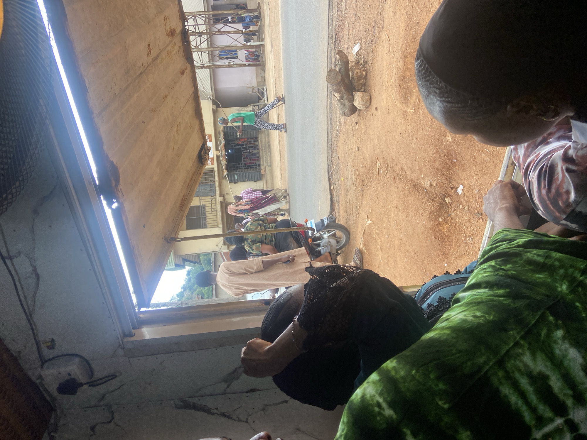 Captured from an interior perspective, the image presents a view of a street scene in Okengwe-Obehira, Nigeria. Several individuals are visible along the street, and at least one motorcycle is present. The roadside exhibits dirt and some wood debris. Inside the structure from which the image is taken, multiple people are partially visible. The interior structure appears weathered.