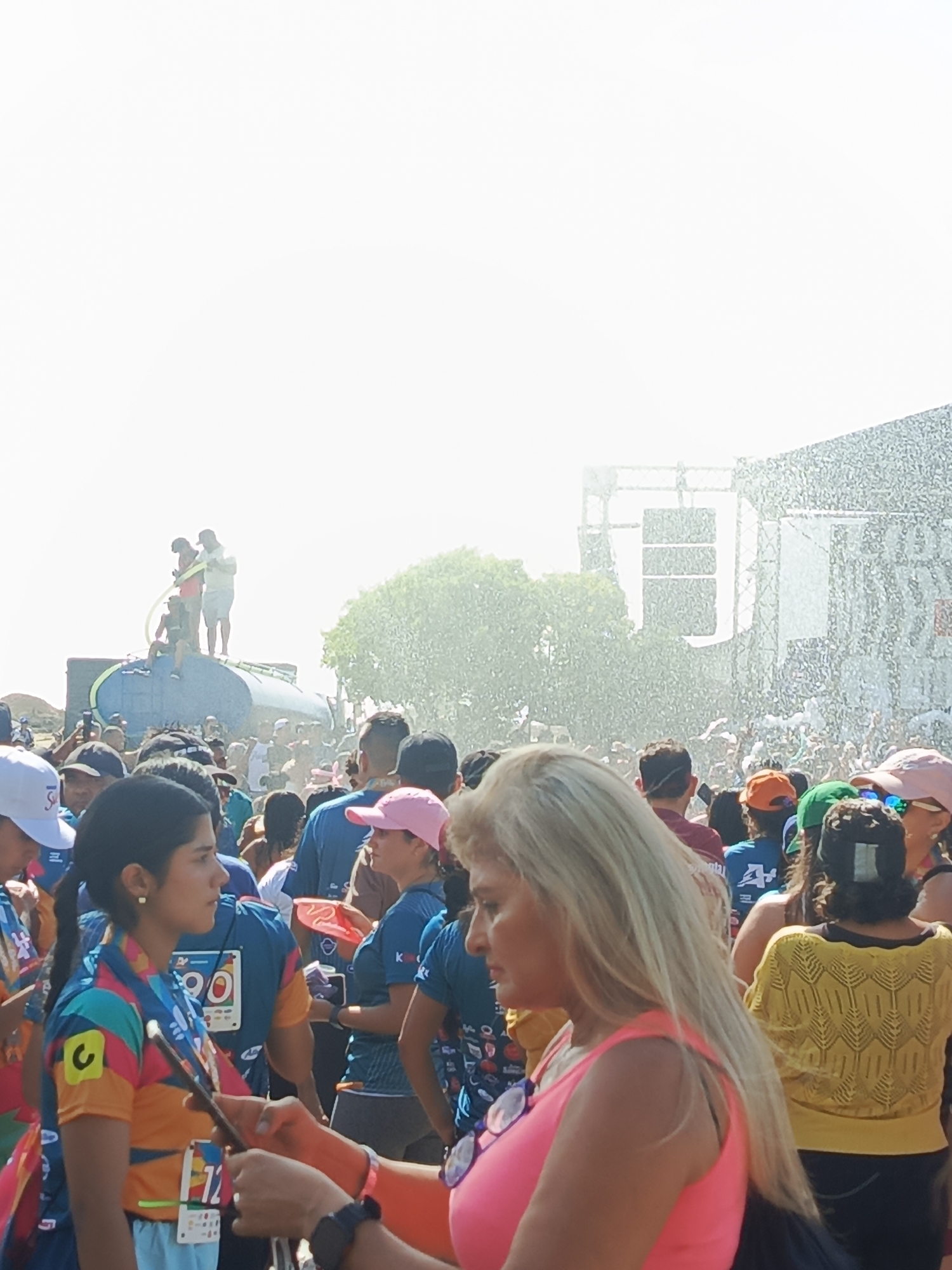 The image captures a densely crowded outdoor event in Porlamar, Venezuela, on a bright, sun-drenched day. In the foreground on the right, a blonde woman in a pink tank top is partially visible, seen in profile looking towards the left, with sunglasses hanging from her top. Adjacent to her on the left, a woman with dark hair, wearing a colorful athletic shirt (teal, orange, pink, and yellow) with a green and white race bib numbered "12" and a lanyard, is also seen in profile, looking to the right.
The midground is filled with numerous individuals wearing various casual and athletic attire, including hats. Towards the center-left, several people are standing atop a large, blue, horizontal water tanker or float. At least two individuals on the tanker are actively operating a hose, spraying water over the surrounding crowd, which is partially obscured by the spray. Behind the tanker, green foliage from trees is visible.
In the right background, a large, tall, metallic-looking structure, possibly a stage or scaffolding, is prominently featured. From this structure, a significant volume of white liquid or particulate matter, consistent with water or foam, is being forcefully ejected, creating a dense, misty effect that partially obscures the area behind it and extends over a portion of the crowd. The sky above is largely overexposed and bright white.