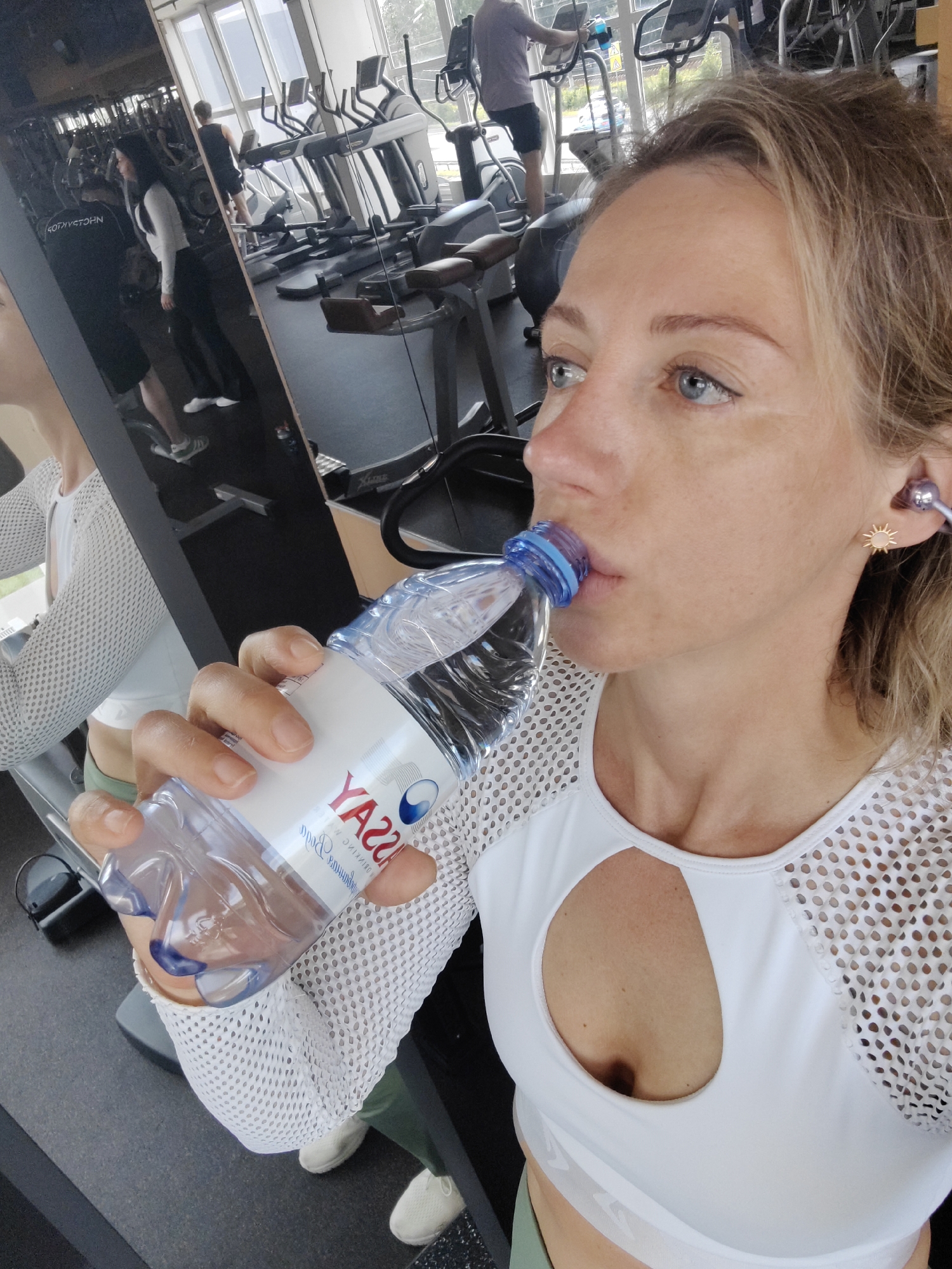 A woman with blonde hair and blue eyes takes a selfie in a gym, drinking from a clear plastic bottle of water. She is wearing a white mesh long-sleeved top with a cutout revealing her chest, and a white sports bra with a logo. She also has on olive green leggings and white sneakers. In the background, reflections in a large mirror show other gym equipment, including treadmills and elliptical machines, with a few other people working out. The setting appears to be an indoor fitness center, with large windows providing natural light. The woman's expression is neutral, and she is focused on drinking. The water bottle label is partially visible and reads "Yassa" in large red lettering with a blue circular logo. The overall impression is a candid moment of hydration during a workout.
