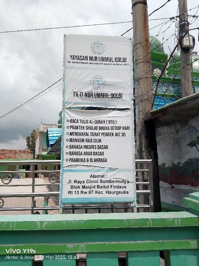 A white sign with blue and red lettering hangs on a metal fence post in front of a green building in Kecamatan Haurgeulis, Indonesia. The sign reads, “Yayasan Nur Ummul Qolbi” at the top and “TK IT Nur Ummul Qolbi” below. There are six bullet points on the sign, outlining the school’s curriculum, which includes “Baca Tulis Al-Quran (BTQ),” “Praktek Sholat Dhuha Setiap Hari,” “Menghafal Surat Pendek Juz 30,” “Manasik Haji Cilik,” “Bahasa Inggris Dasar,” “Bahasa Arab Dasar,” and “Pramuka & Olahraga.” The sign concludes with the school’s address: “Alamat: Jl. Raya Cinini Sumbermulya Blok Masjid Baitul Firdaus Rt 13 Rw 07 Kec. Haurgeulis”. The green building behind the sign appears to be a mosque or place of worship. A portion of the sign is obscured by a metal fence that runs across the front of the building. The sign is being photographed with a mobile phone held in portrait mode. The date and time, “23 Mar 2023, 12:43,” appears in the lower right corner of the image. The image is taken from a low angle, pointing upwards towards the sign.