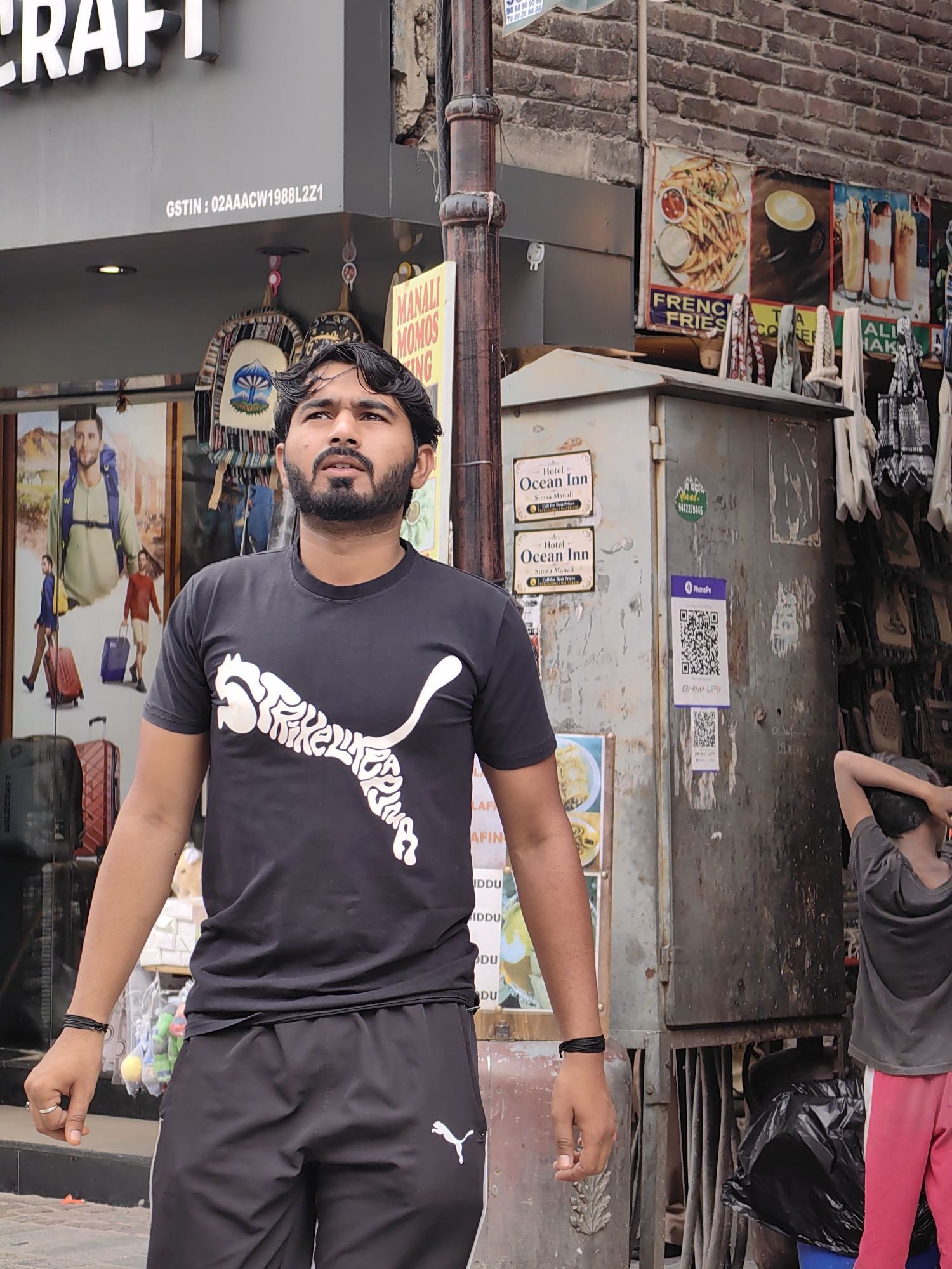 The image depicts a street scene in Manali, India, likely near a market or commercial area. In the foreground, a young man with a beard is looking upwards and to his right. He is wearing a black t-shirt with a white Puma logo made of intricate white lettering, and black athletic pants with a white stripe and a small white Puma logo. He has a black bracelet on his left wrist and a ring on his right index finger. Behind him, a poster advertises "MANALI MOMOS KING".
To the left of the young man, a large poster features a man with a backpack and other figures carrying luggage. Various bags and suitcases are visible in the lower left of the frame.
To the right of the young man, there is a large, weathered metal electrical box or utility cabinet. Two signs for "Hotel Ocean Inn" in Simla Manali are affixed to the cabinet. A purple sticker with a QR code and "PhonePe" is also present. Further to the right, a younger person in a grey t-shirt and bright pink pants is standing with their back to the camera, their right arm raised to their head. In the background, posters displaying food items like french fries and drinks, as well as various textiles and hanging items, are visible on a brick wall. A black trash bag and a blue bucket are present in the lower right corner. The environment appears to be an outdoor setting on a street with visible infrastructure like a lamppost and electrical conduits.