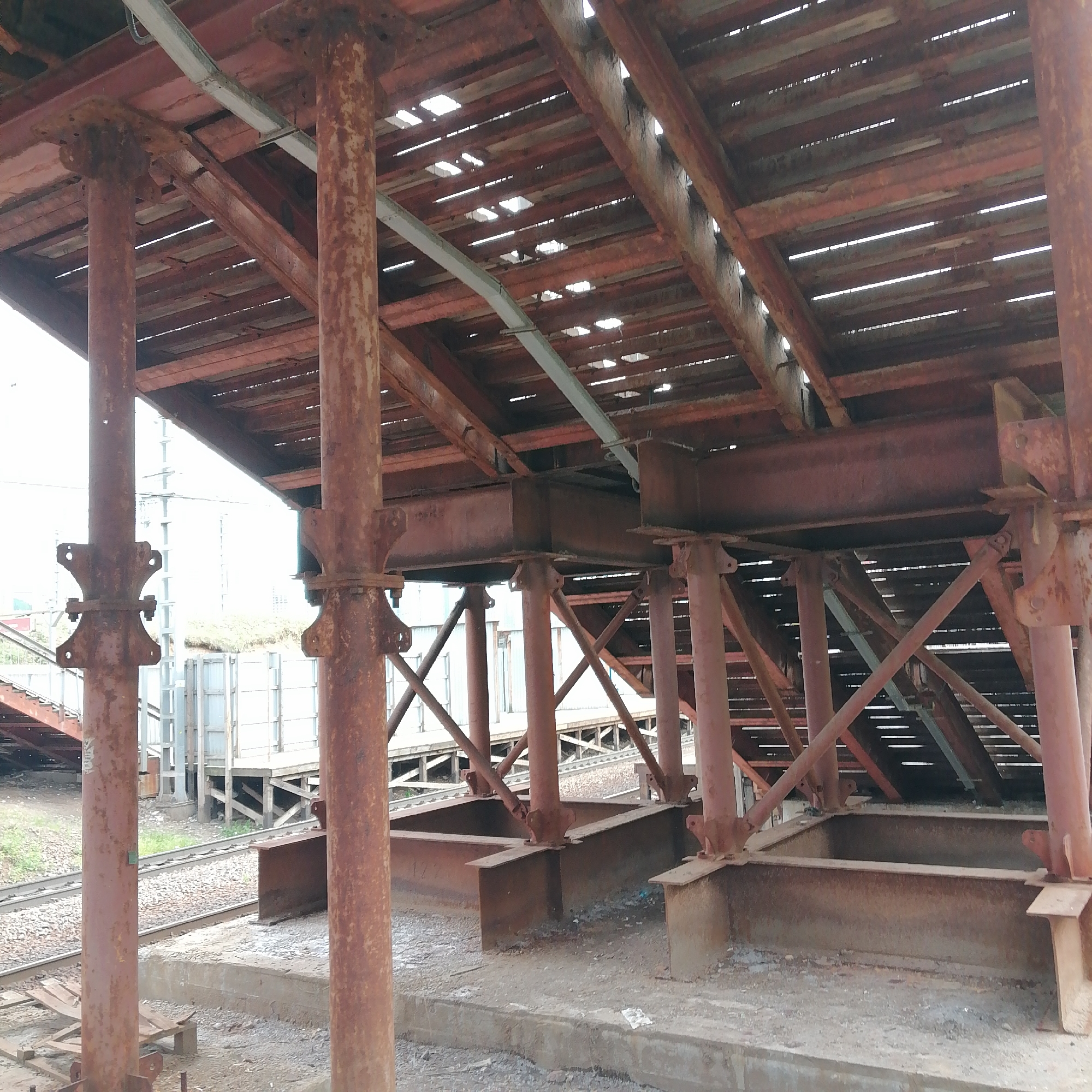 This image captures a heavily rusted, multi-level industrial structure, possibly a loading platform or an elevated walkway, set against a bright, overcast sky during daytime. The primary subjects are the robust, reddish-brown metal components that form the support system and the platform itself.
Numerous thick, cylindrical metal columns, heavily marked with rust, serve as vertical supports. These columns feature bolted connection plates. Spanning between these supports are large, rusted metal beams, some appearing as I-beams and others as box beams. Above these beams, a deck is formed by widely spaced wooden planks, allowing significant light to filter through the gaps. Diagonal metal braces are visible, reinforcing the structure.
At the base, the columns are anchored into substantial concrete or metal foundations, some of which form open, rectangular basin-like structures on the ground. The ground immediately beneath the structure is a mix of gravel and concrete.
In the background, to the left and center, parallel railway tracks are visible, lined with gravel. Further back, there are indistinct industrial buildings or structures and some sparse vegetation. No people are present, and no activities are taking place. The pervasive rust suggests the structure is old, exposed to the elements, or possibly disused. No legible text is visible in the image.