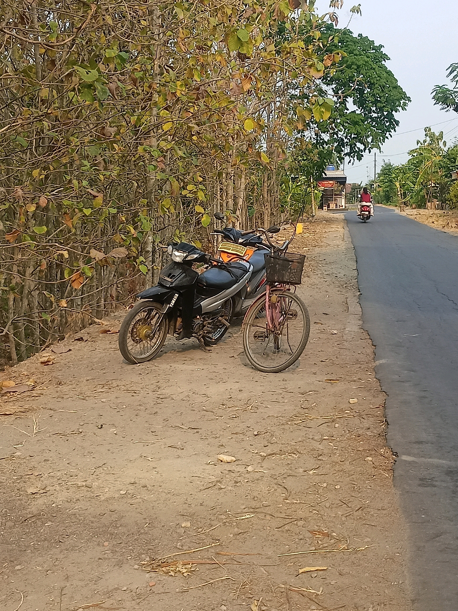 Two motorcycles and a bicycle are parked on a dirt roadside in Sambiduwur, Indonesia. A black motorcycle with silver accents is in the foreground, followed by a pink bicycle with a black basket. Behind these vehicles, a dirt path runs alongside a paved road. A person on a red motorcycle is traveling away from the viewer on the road. Trees and foliage line the left side of the road, with a small structure visible in the distance. The sky is overcast.