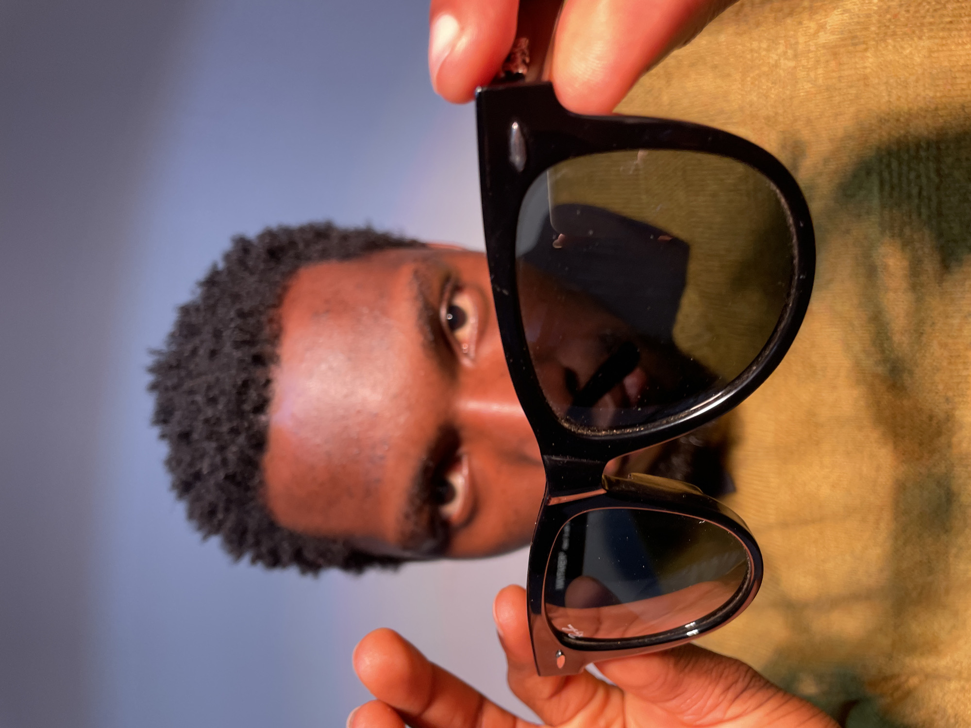 A man of African descent is holding a pair of black sunglasses in front of his face. He has short, dark curly hair and dark brown eyes. His skin tone is dark brown. The sunglasses are black and rectangular with rounded edges. The lenses appear dark. His hands are holding the sunglasses by the frame. The background is blurred, showing a light blue on the left and a muted yellow-green on the right. There is no visible information to determine the location in Barakin Akawo, Nigeria.