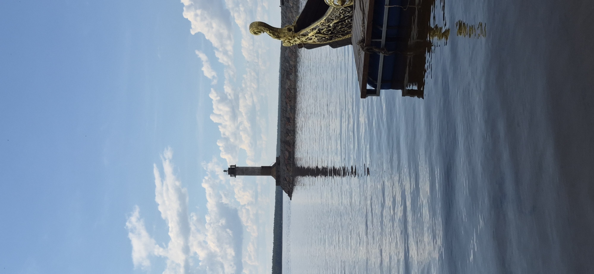 In the city of Motornoe, Russia, a lighthouse stands on a stone pier against a sky filled with scattered white clouds and blue hues. The water is calm and reflects the lighthouse. In the foreground, the ornate golden prow of a boat is visible, tethered to a wooden dock. The scene captures a tranquil waterscape with a historical maritime element.