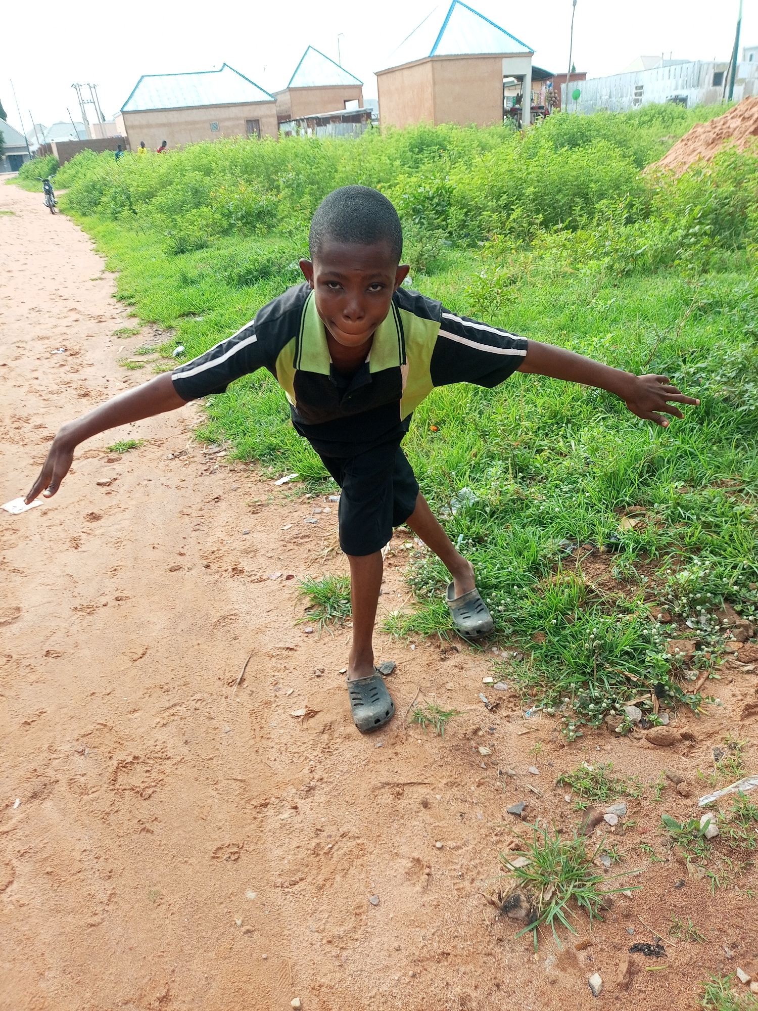 A young Black boy stands on a dirt path, his arms outstretched to the sides as if balancing. He is wearing a black polo shirt with light green accents on the collar and sleeves, and black shorts. His feet are clad in dark, clunky sandals. The boy is looking directly at the camera with a serious expression.
Behind the boy, a lush green area of tall grass and small plants stretches out. In the background, several simple buildings with light-colored walls and metallic blue roofs are visible, suggesting a rural or semi-urban setting. The overall environment appears to be outdoors, possibly in the city of Tempure, Nigeria, given the style of the buildings and the vegetation. The path the boy is standing on is made of reddish-brown dirt.