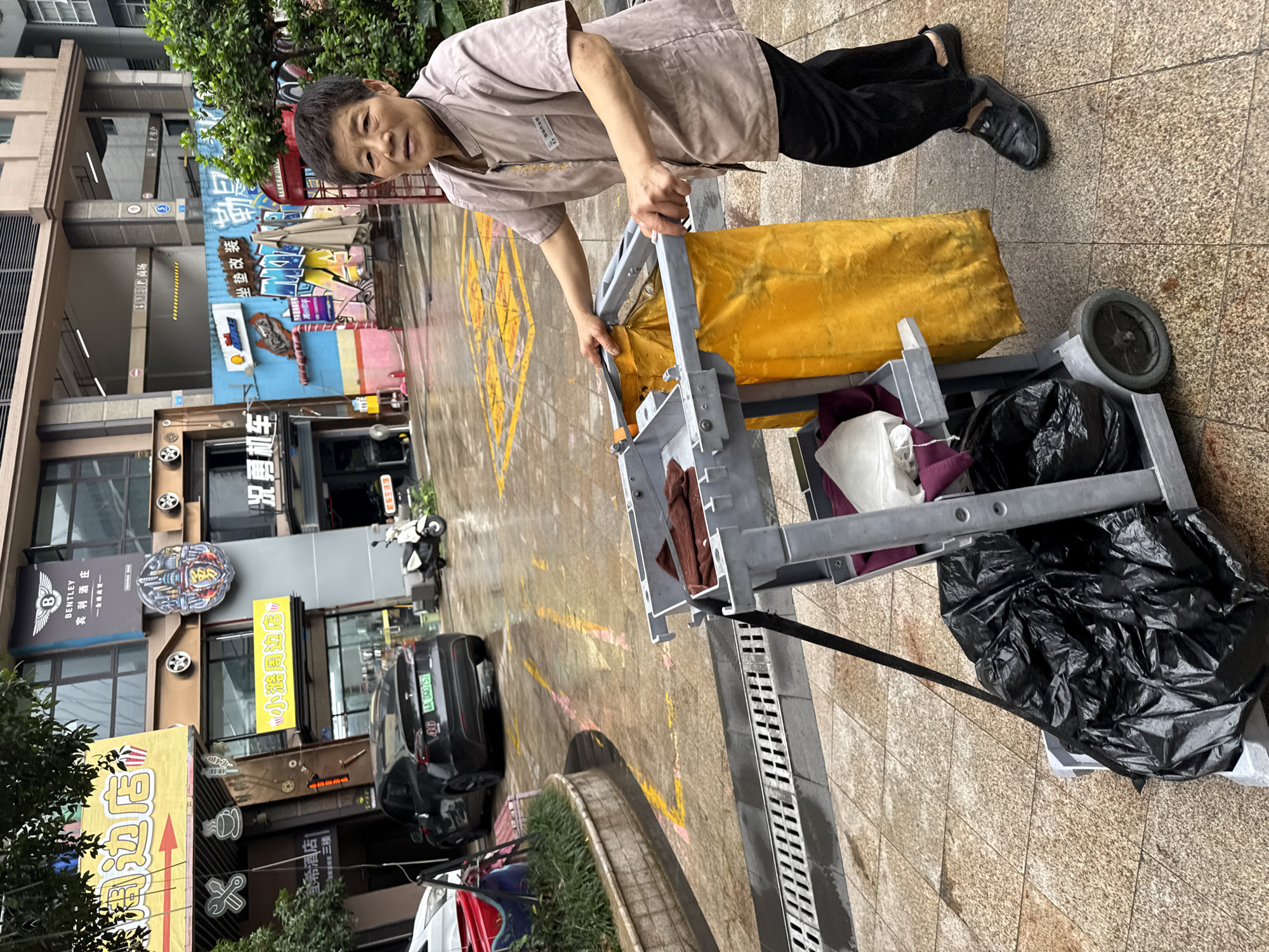 Here is a detailed description of the image:
**Scene:** The image captures a street scene in what appears to be the Nan'an District of China. This is indicated by the license plate visible in the image. The area is urban, with buildings on either side of the street.
**Person:** The primary subject is an adult Asian individual. They are dressed in a brown button-up shirt and black pants, likely a uniform. The individual is standing and appears to be pushing or maneuvering a cleaning cart.
**Objects:**
* Cleaning Cart: The cart is loaded with what seems to be cleaning supplies. There is a large yellow bag, likely for collecting waste. Black trash bags are also visible.
* Buildings and Shops: The buildings have storefronts, including a Bentley dealership and a shop with auto-related signage.
* Vehicles: Cars and scooters are parked on the street, adding to the urban environment.
**Actions/Interactions:** The person is actively engaged in cleaning or maintenance work. The individual appears to be aware of the camera, as there is some eye contact.
**Environment:** The setting seems to be an outdoor area, potentially a pedestrian zone or a street with shops. There are drainages in the pavement.