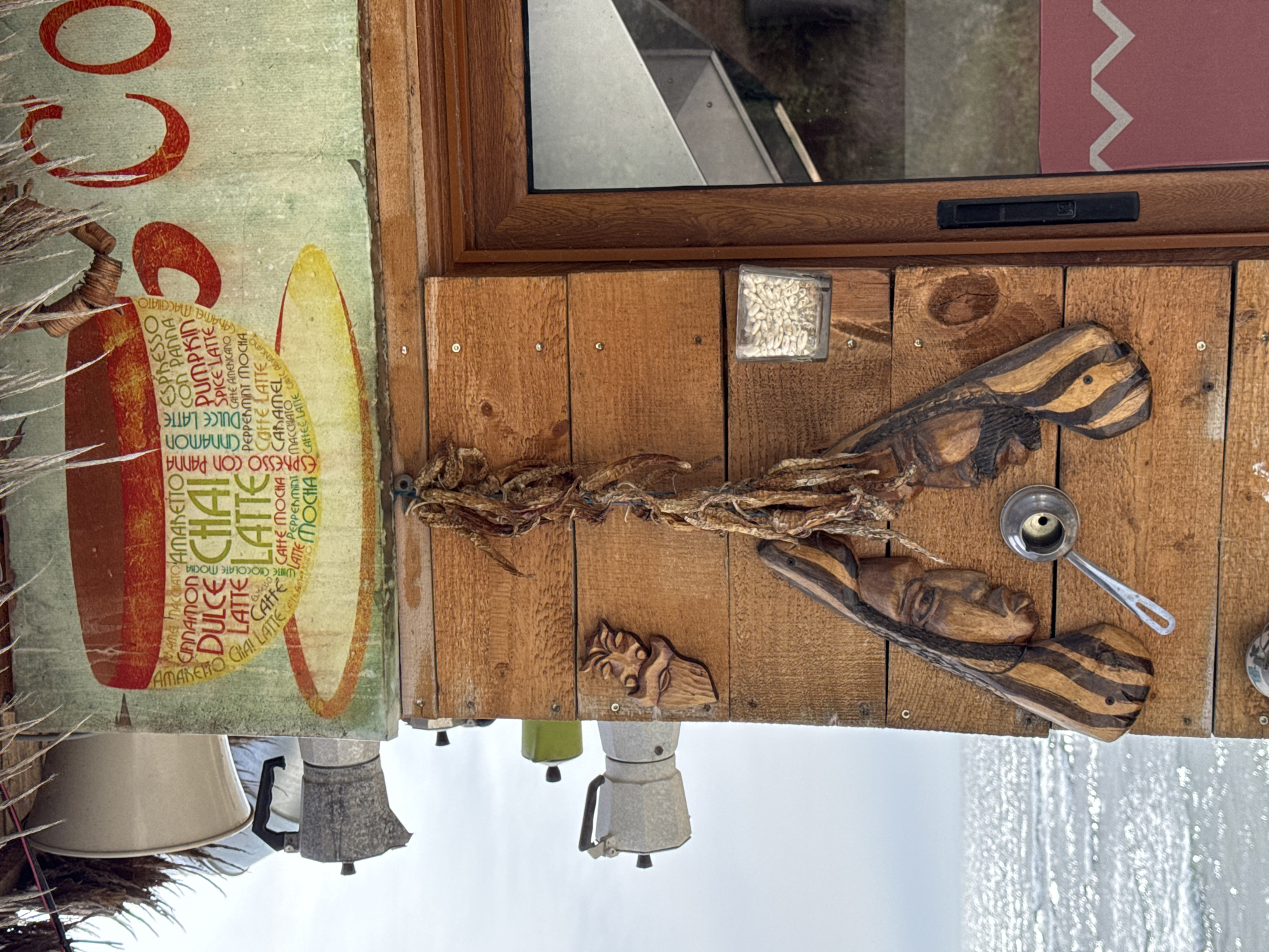 The image depicts a detail of what appears to be an outdoor coffee stand or small shop, possibly located in Varna, Bulgaria (though this is inferred without direct visual confirmation of the city). The scene is shot close-up, focusing on the exterior facade.
A portion of a sign with coffee-related terms like "Cinnamon", "Pumpkin Spice Latte", "Dulce Latte", "Chai Latte", and "Mocha" is visible, suggesting the shop's primary offering.
Attached to the wooden facade of the structure is a collection of decorative items. These include two wooden carvings of faces, a small metal scoop, and what appears to be a string of dried peppers or herbs. Above, there is a window with a partially visible interior and a red-patterned awning above.
At the bottom of the frame, a few Moka pots are suspended as decoration, indicating an additional focus on coffee preparation. The background shows the sky, suggesting an outdoor setting.