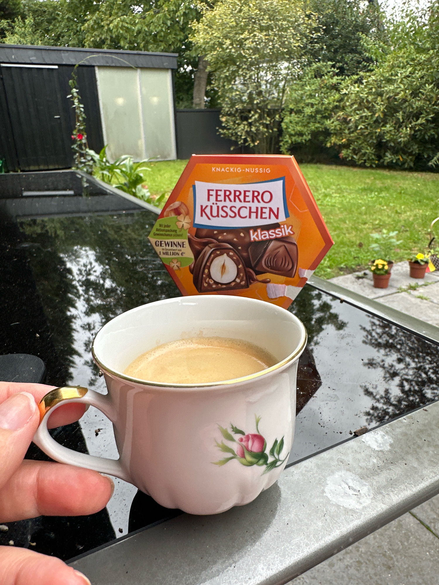 A hand holds a pink and gold rimmed ceramic coffee cup filled with coffee, positioned on a black reflective surface, possibly a table, outdoors. Behind the cup is a hexagonal orange box of Ferrero Küsschen chocolates, featuring images of the chocolates and text in German. The background reveals a garden with lush green trees, a dark wooden shed with a translucent panel, and a paved area with small potted plants. The overall scene suggests a moment of relaxation in a residential outdoor setting in Krefeld, Germany, as indicated by the presence of Ferrero Küsschen, a popular brand in Germany.