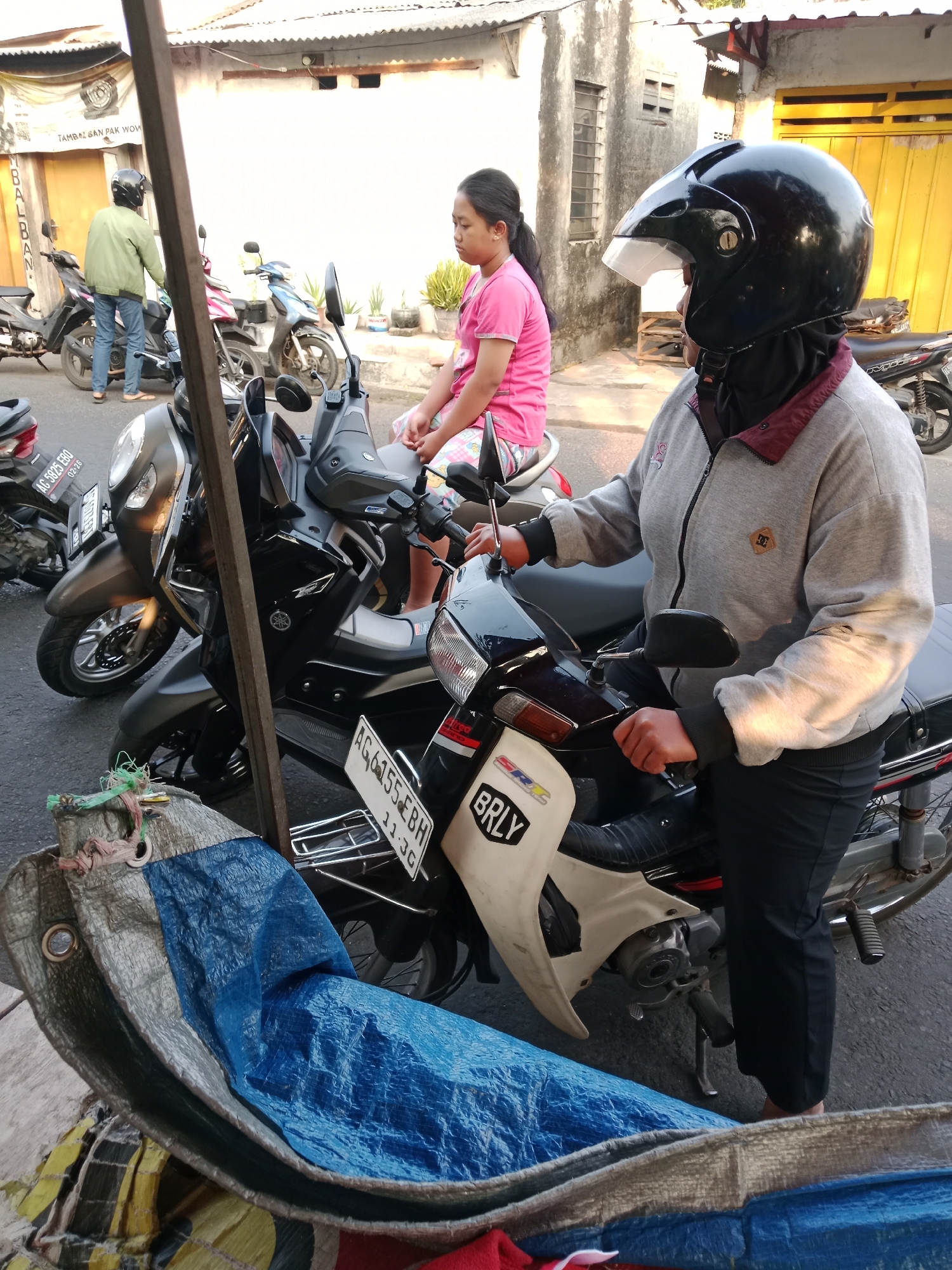 In Kediri City, Indonesia, a young Indonesian girl wearing a pink t-shirt and patterned shorts sits on a motorcycle. Beside her, a person in a grey zippered jacket and black pants, wearing a black helmet and a black face covering, is also seated on a motorcycle. The motorcycles are parked on a paved street. In the foreground, a blue tarp is draped over some objects, and in the background, a building with a yellow door and a tiled roof is visible, with a white wall and some windows. The scene appears to be outdoors, possibly on a street or in a parking area.