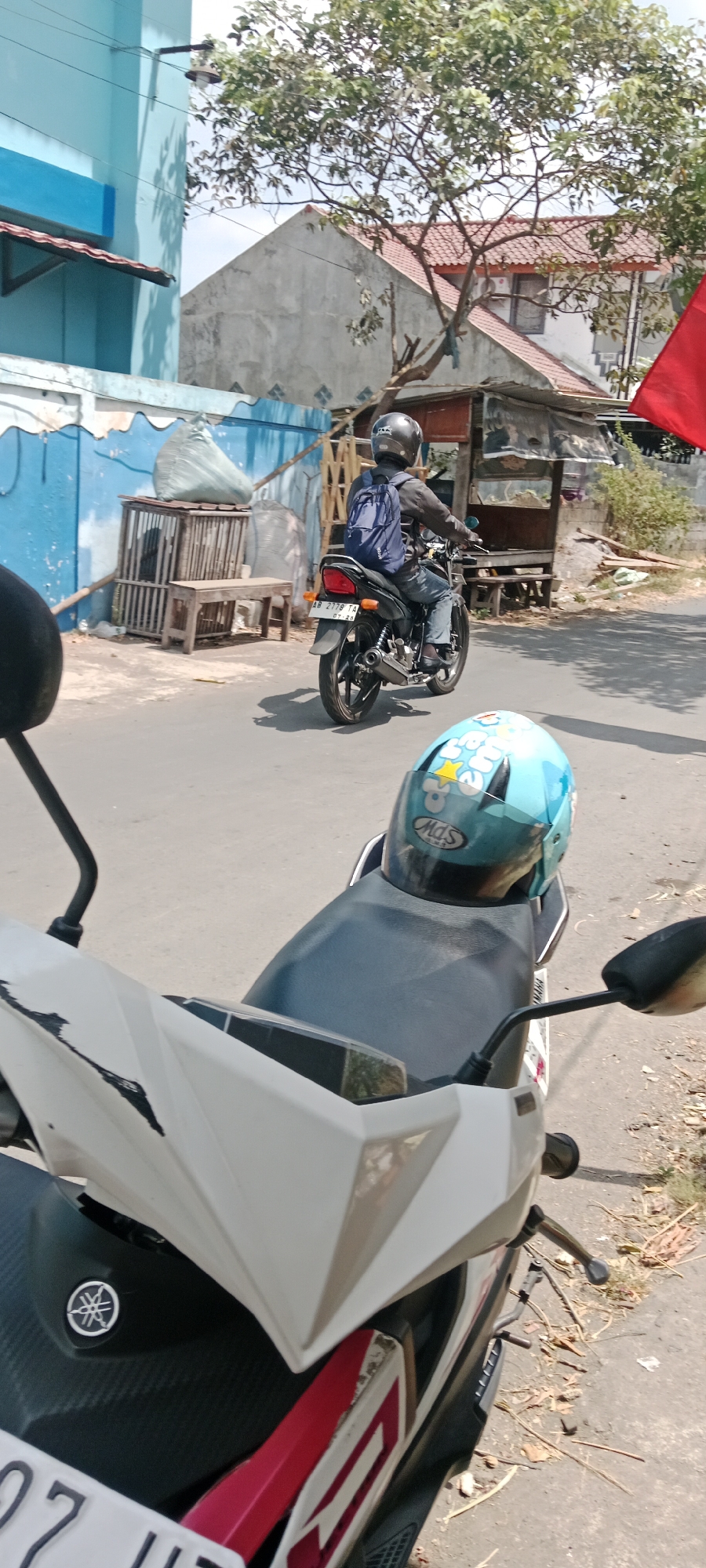In the city of Purwomartani, Indonesia, a person is riding a black motorcycle down a paved road. The rider is wearing a black helmet and a blue backpack. The motorcycle has a license plate that reads "AB 2779 TA 07-24". In the foreground, a white motorcycle with a black seat is parked. A light blue helmet with cartoon characters is placed on the seat of the parked motorcycle. The background shows a light blue building with a red awning, a grey building with a red tiled roof, and some trees with green leaves. There is also a wooden cage and a bench next to the blue building. The scene appears to be a street on a sunny day.
